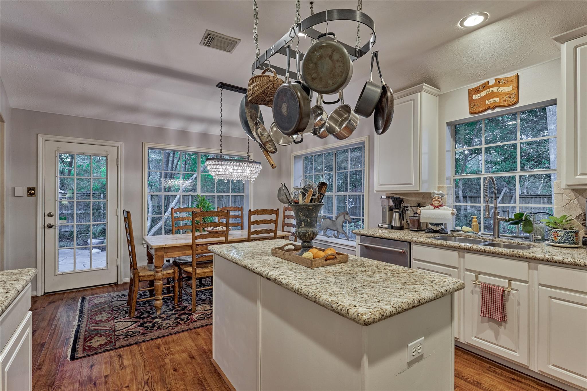 Kitchen with a large granite island, hanging pots and pans, and a dining table beside wooded windows.