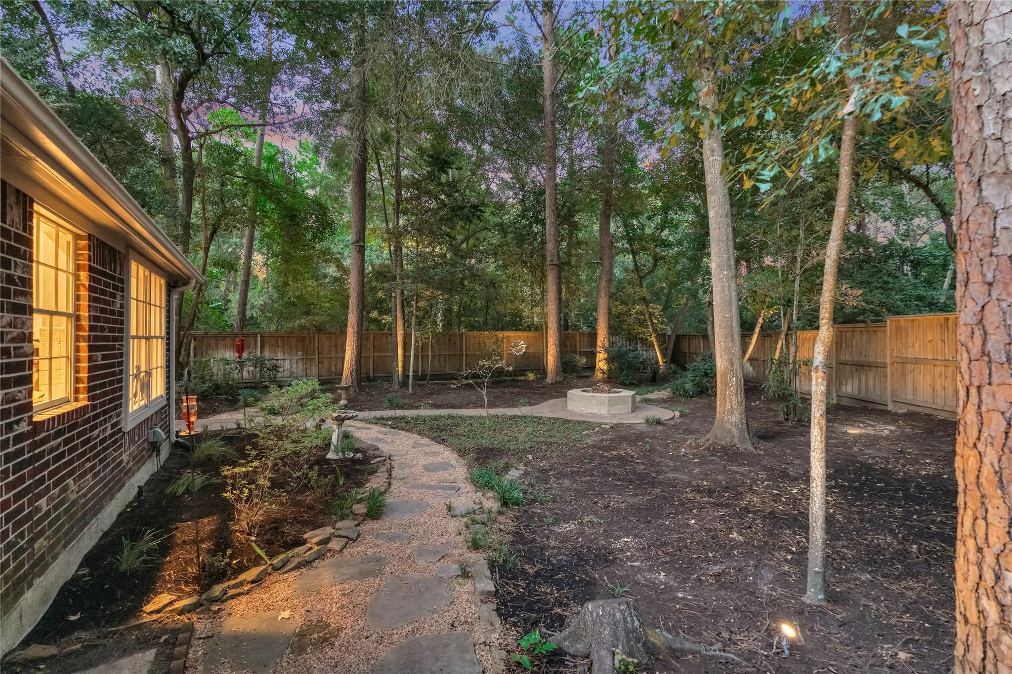 Wooded backyard with a curved stepping-stone path, mature trees, and a fenced yard beside a brick home.