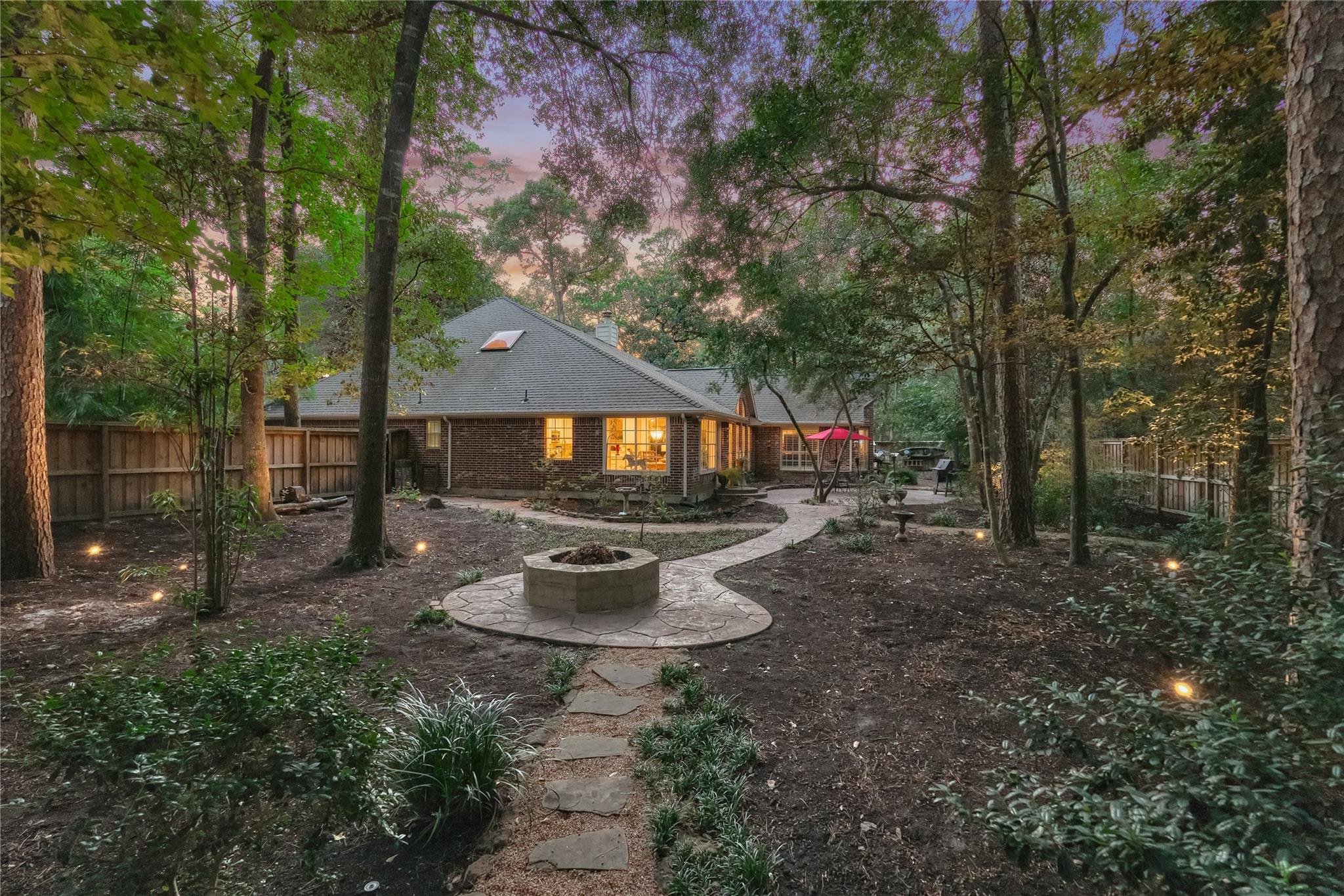 Brick home's wooded backyard with a winding stone path, circular fire pit, and landscape lighting.