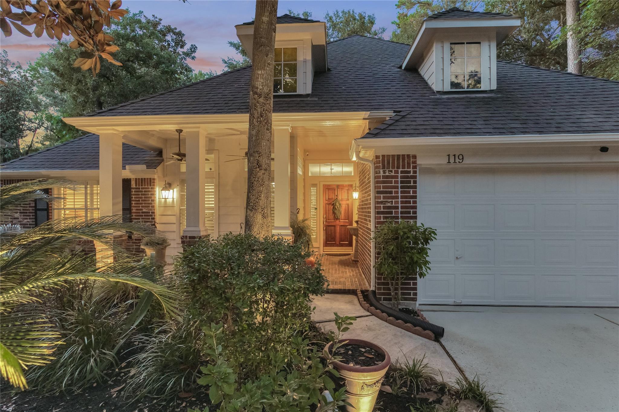 Home exterior at dusk with a warmly lit covered porch, dormer windows, and attached garage.