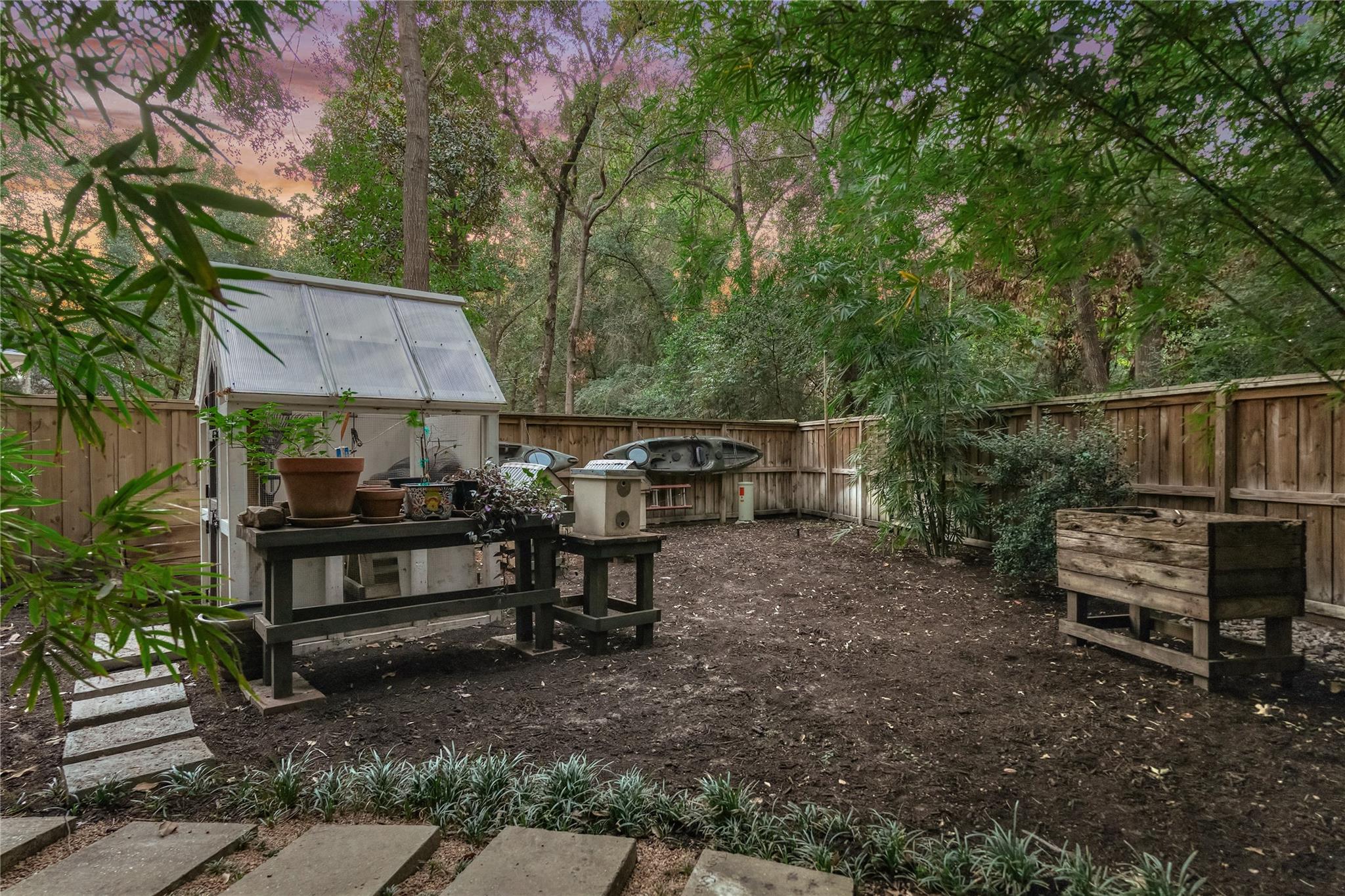 Fenced, wooded backyard with a small greenhouse, planting tables, and kayaks along the fence.