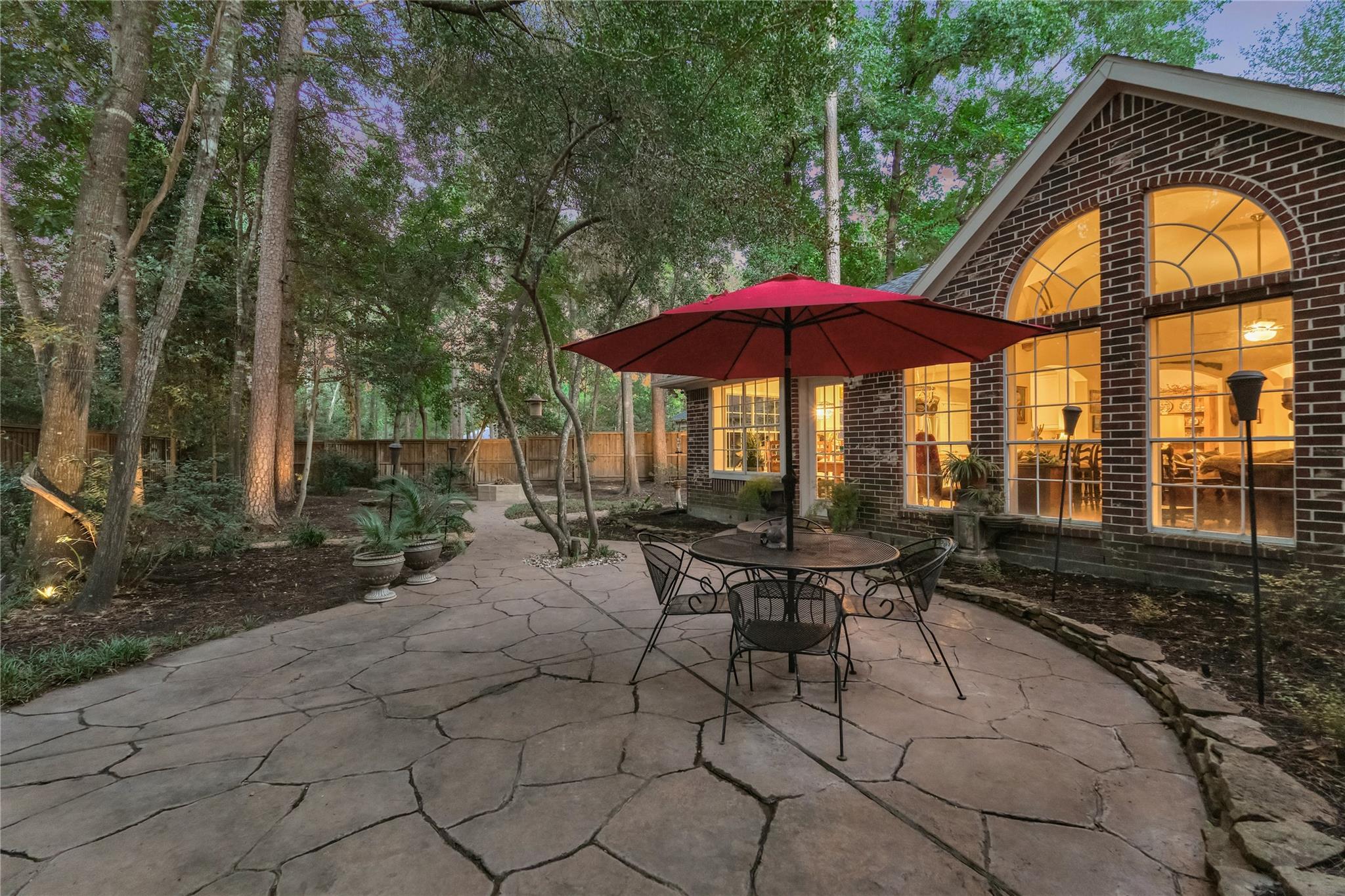 Flagstone patio with a red umbrella dining set beside a brick home with glowing arched windows.