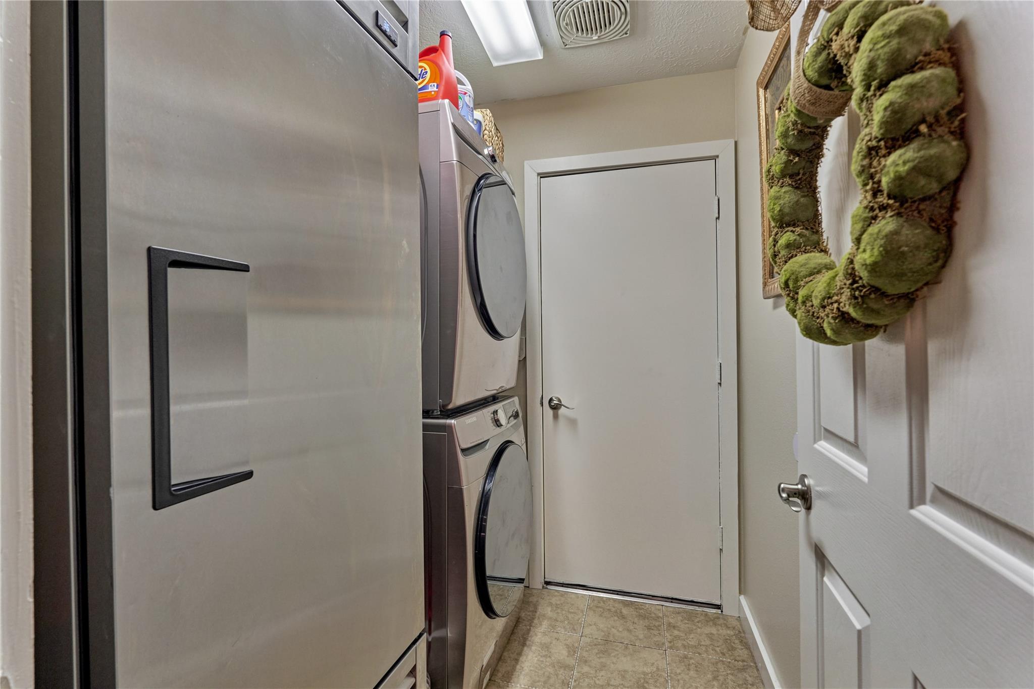 Laundry room with stacked washer and dryer, a stainless refrigerator, and beige tile flooring.