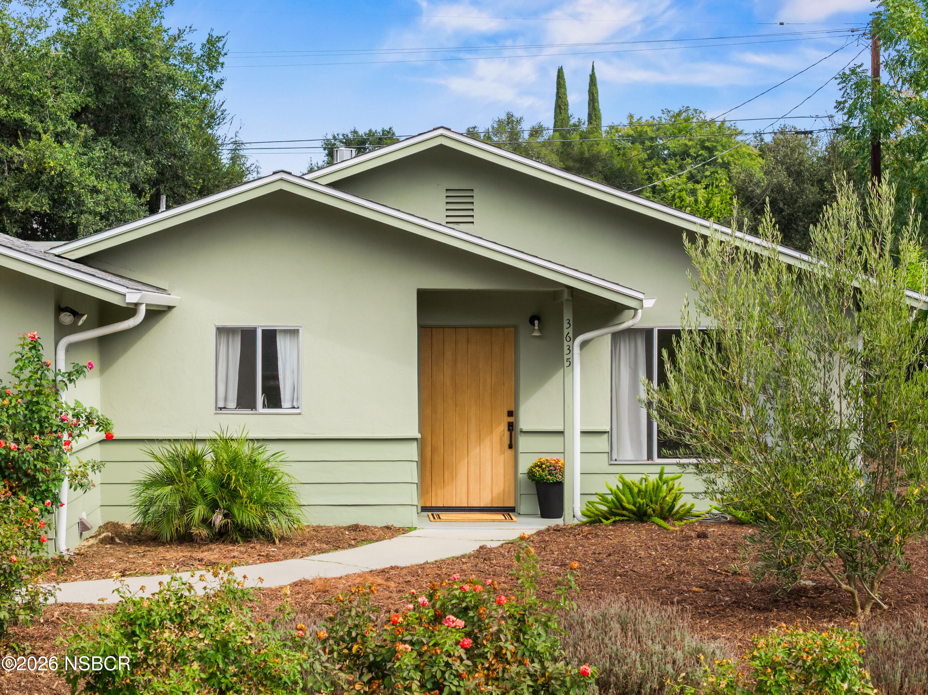 Pale green home with a wood front door, peaked roof, and flowering garden.