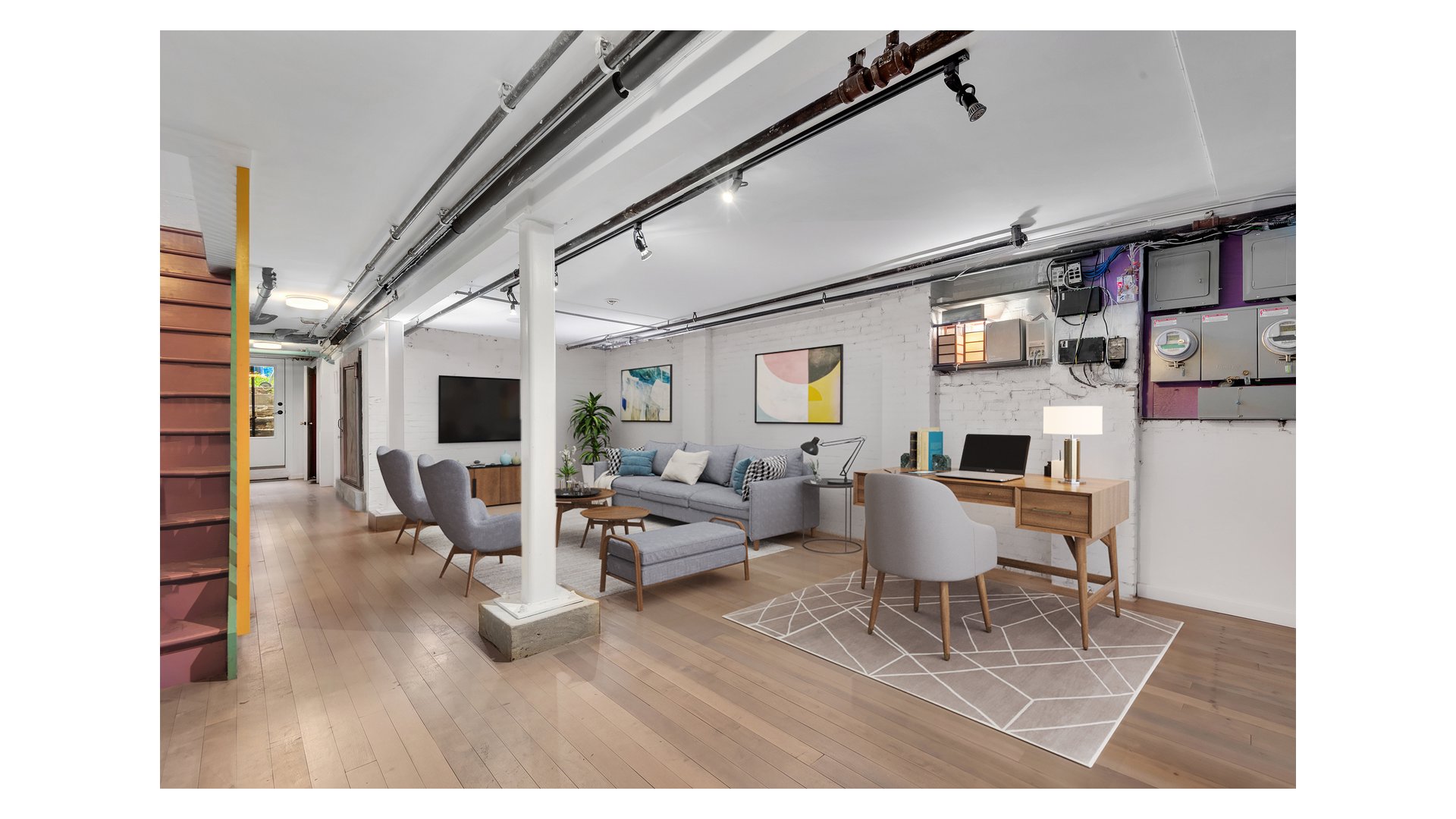 Staged living room with a gray sofa, desk, exposed ceiling pipes, and a staircase.