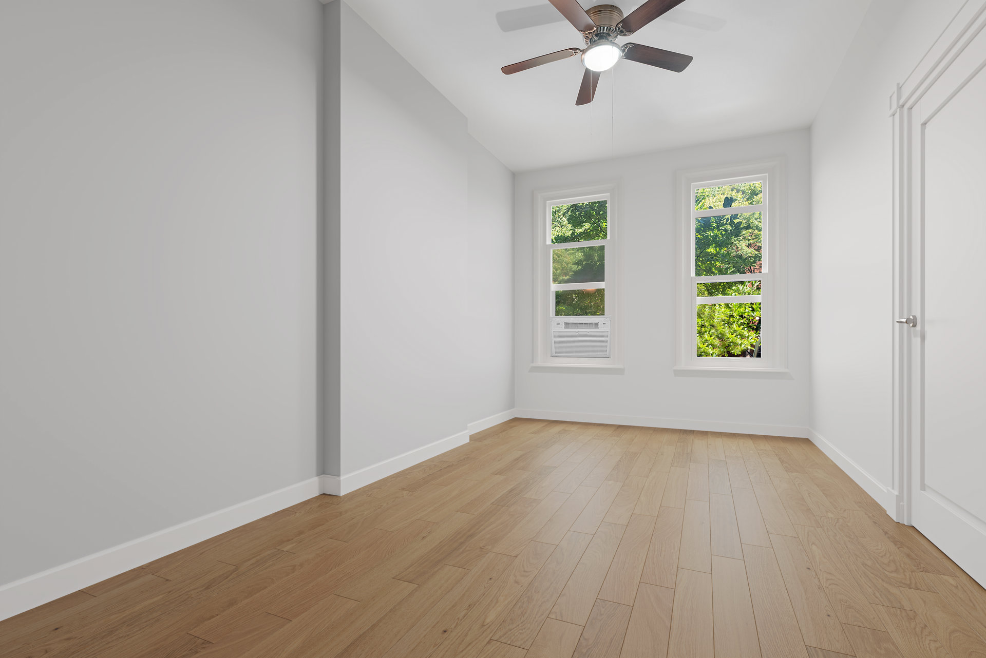 Vacant room with light wood floors, two leafy-view windows, and a ceiling fan.