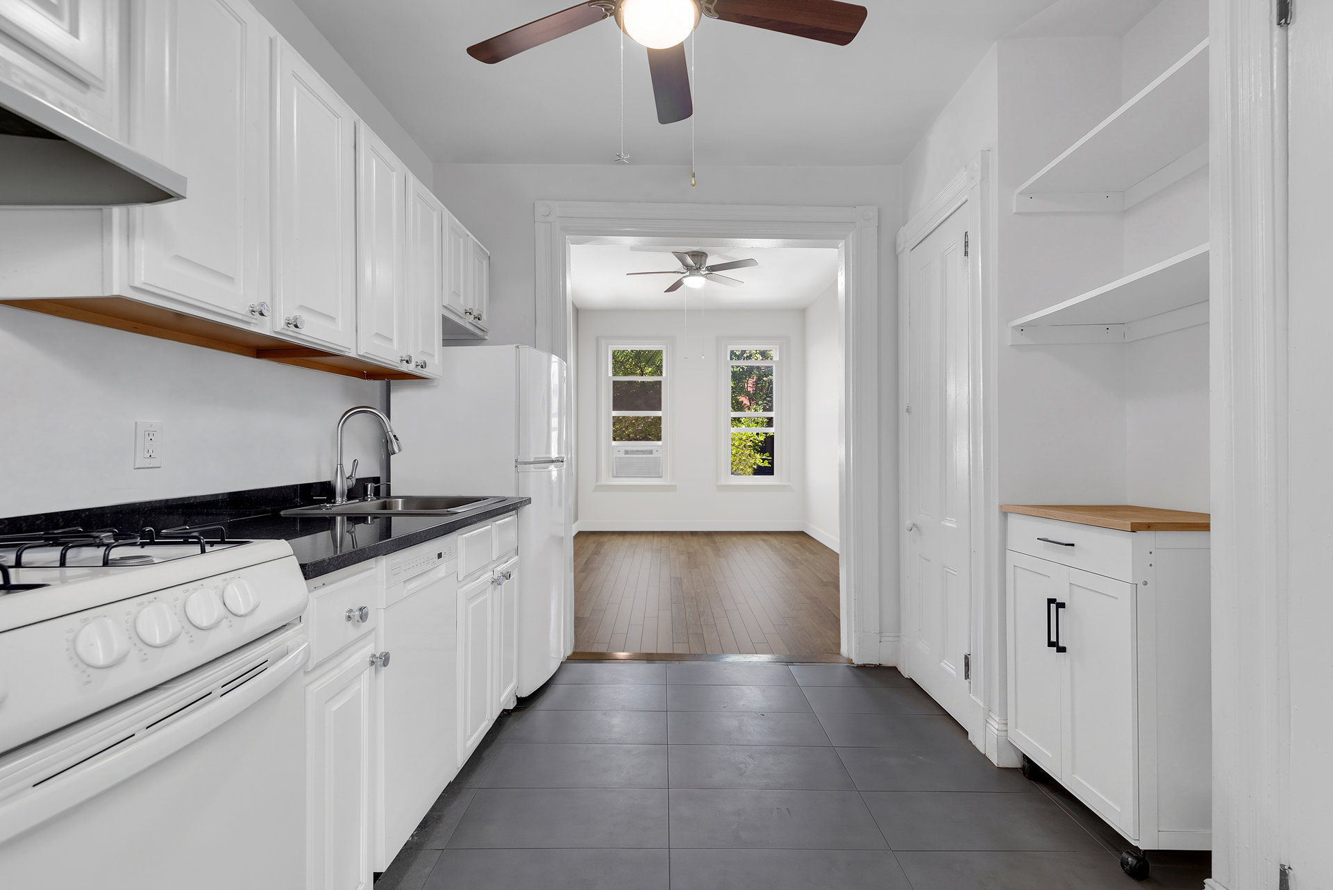 Vacant kitchen with white cabinetry, black countertops, and a view into a sunlit room with hardwood floors.