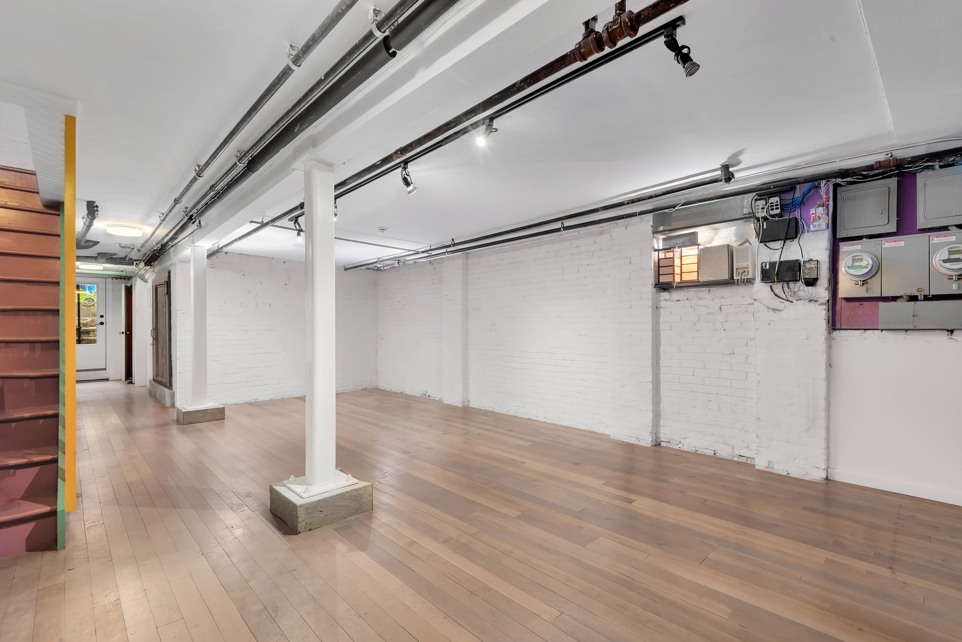 Finished basement with pale wood flooring, exposed ceiling pipes, and a staircase.