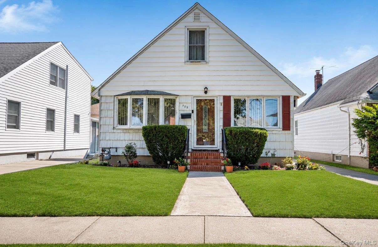 White steep-gabled house with red shutters, brick steps, flower beds, and a broad green lawn.