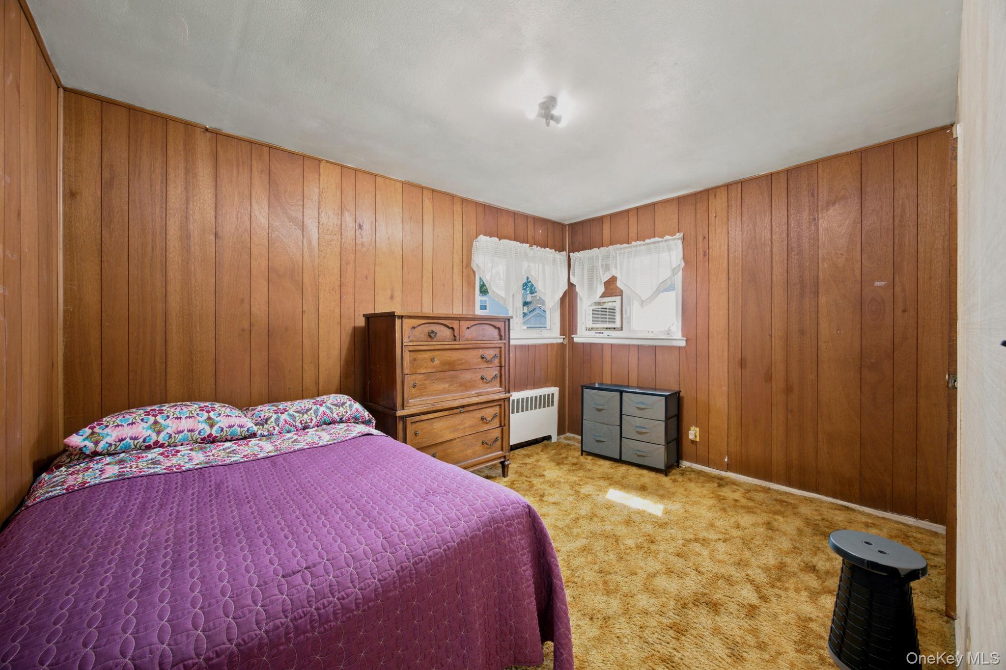 Bedroom with wood-paneled walls, a purple bedspread, wooden dresser, and two curtained windows.