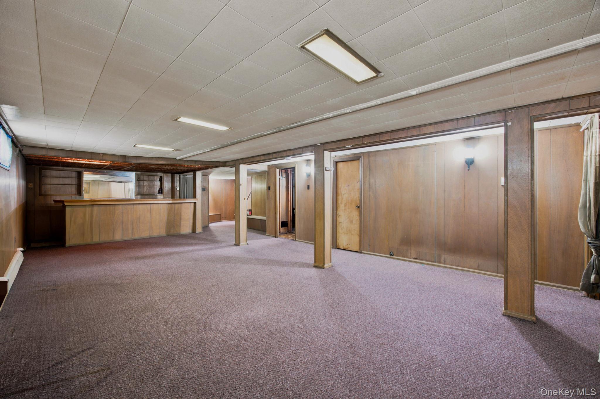 Empty basement room with mauve carpeting, wood paneling, support columns, and a built-in bar.