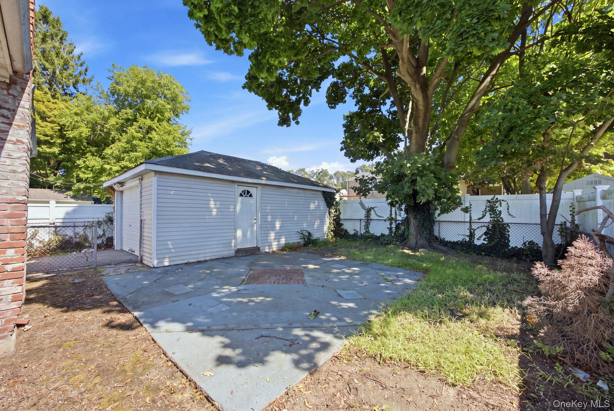 Detached white garage beside a paved patio, with mature trees and fencing.