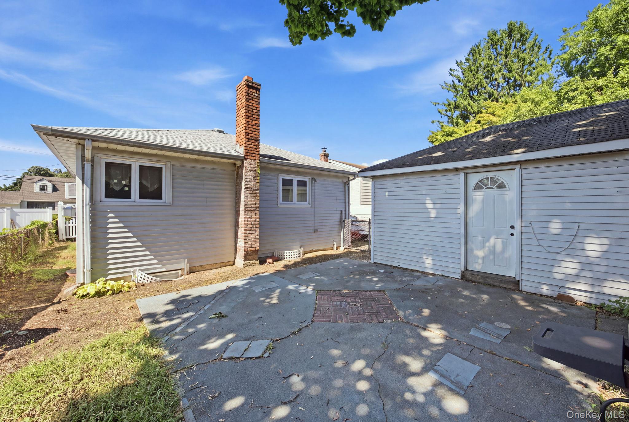Rear of a gray-sided house with a brick chimney, attached garage, and cracked concrete patio.