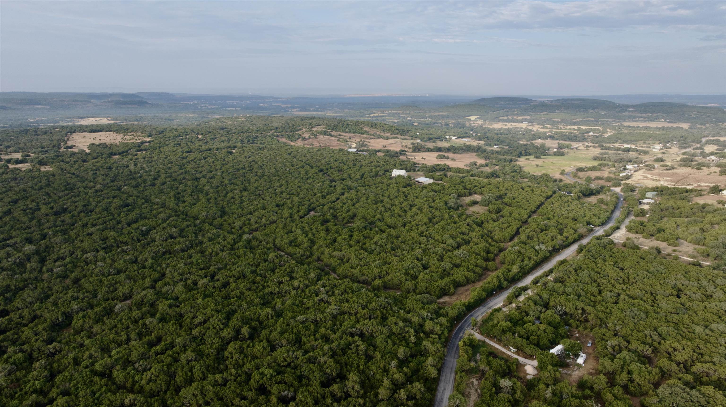 Aerial view of wooded acreage crossed by a winding road, with scattered rural buildings and rolling hills beyond.