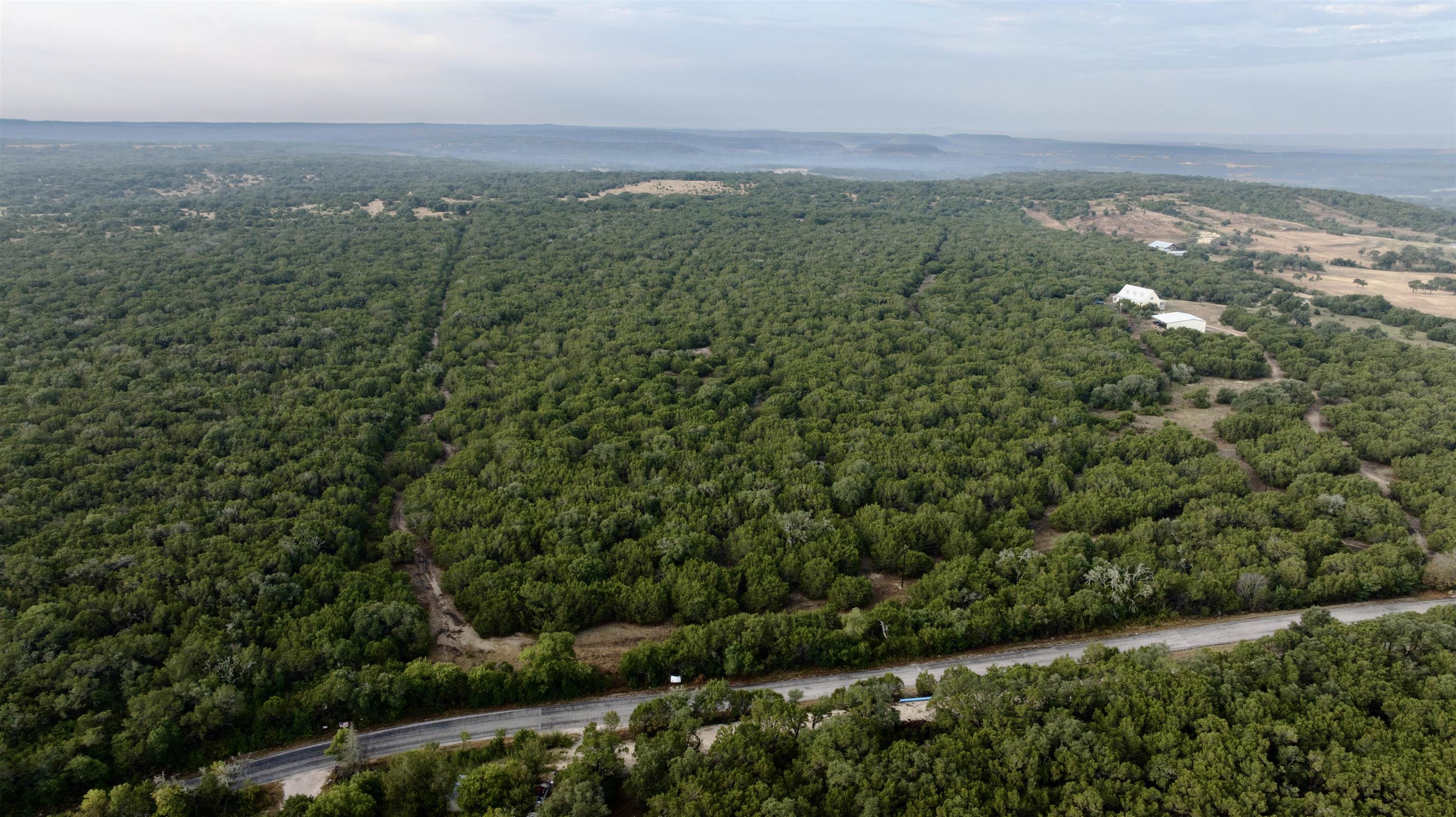 Aerial view of wooded land with road frontage, dirt tracks, and distant buildings.
