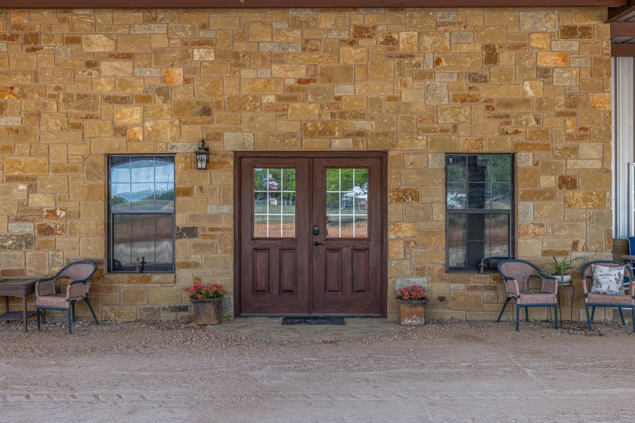 Stone home entrance with dark double doors, flanking windows, porch chairs, and flower pots.