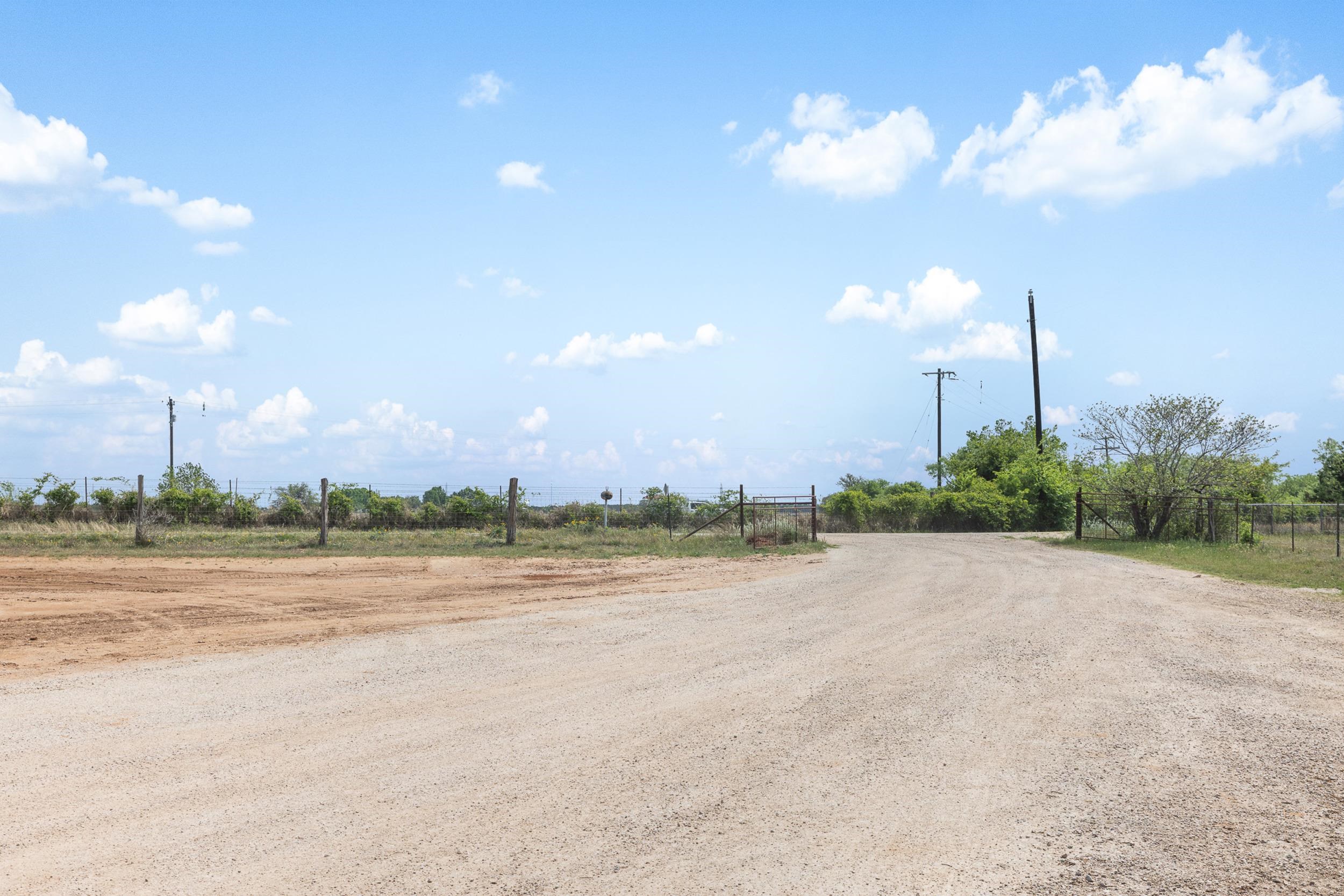 Gravel entrance and metal gates at a fenced rural property under a blue, cloud-filled sky.