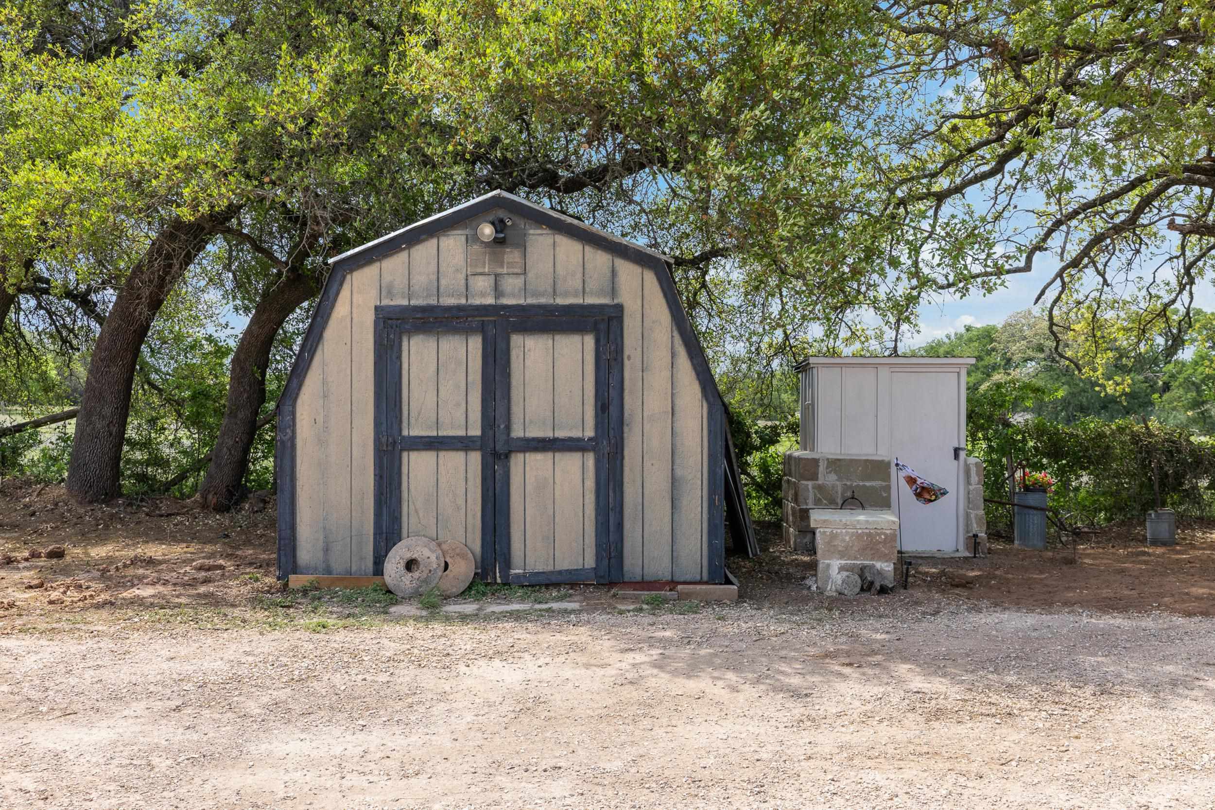 Gray storage shed with dark blue trim beneath leafy trees, beside a smaller shed and stone trough.