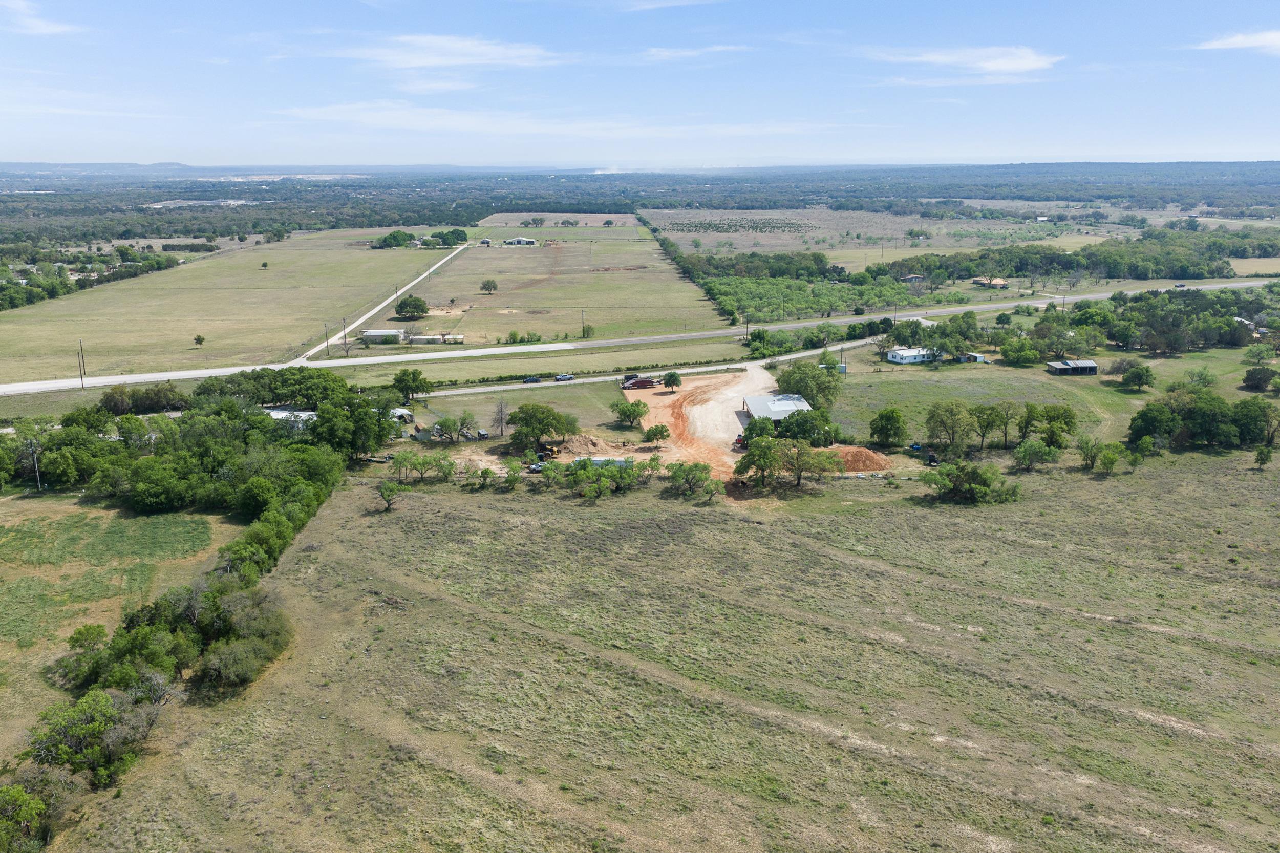 Aerial view of open rural acreage with scattered trees, a cleared dirt area, and a metal-roof building.
