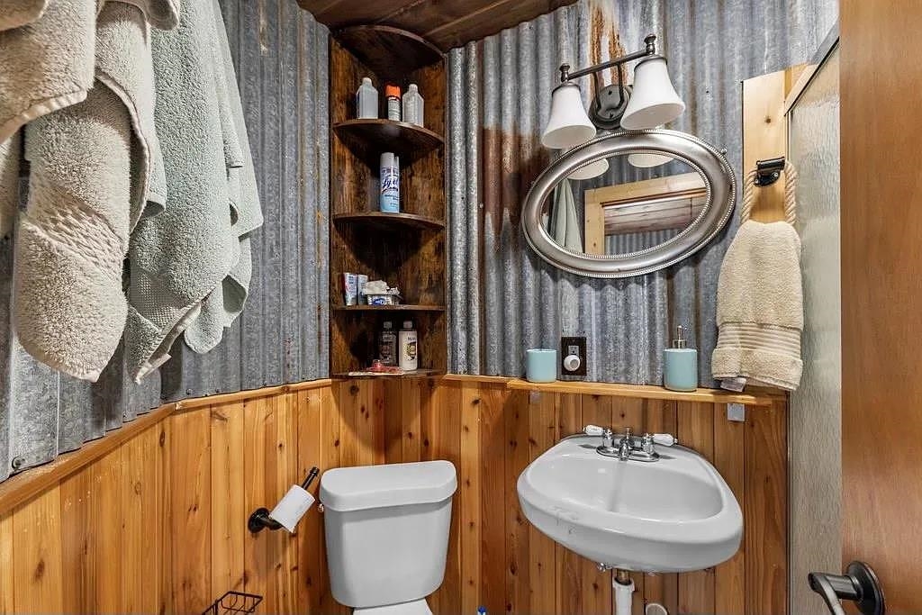 Bathroom with corrugated metal walls, wood paneling, corner shelves, an oval mirror, toilet, and sink.