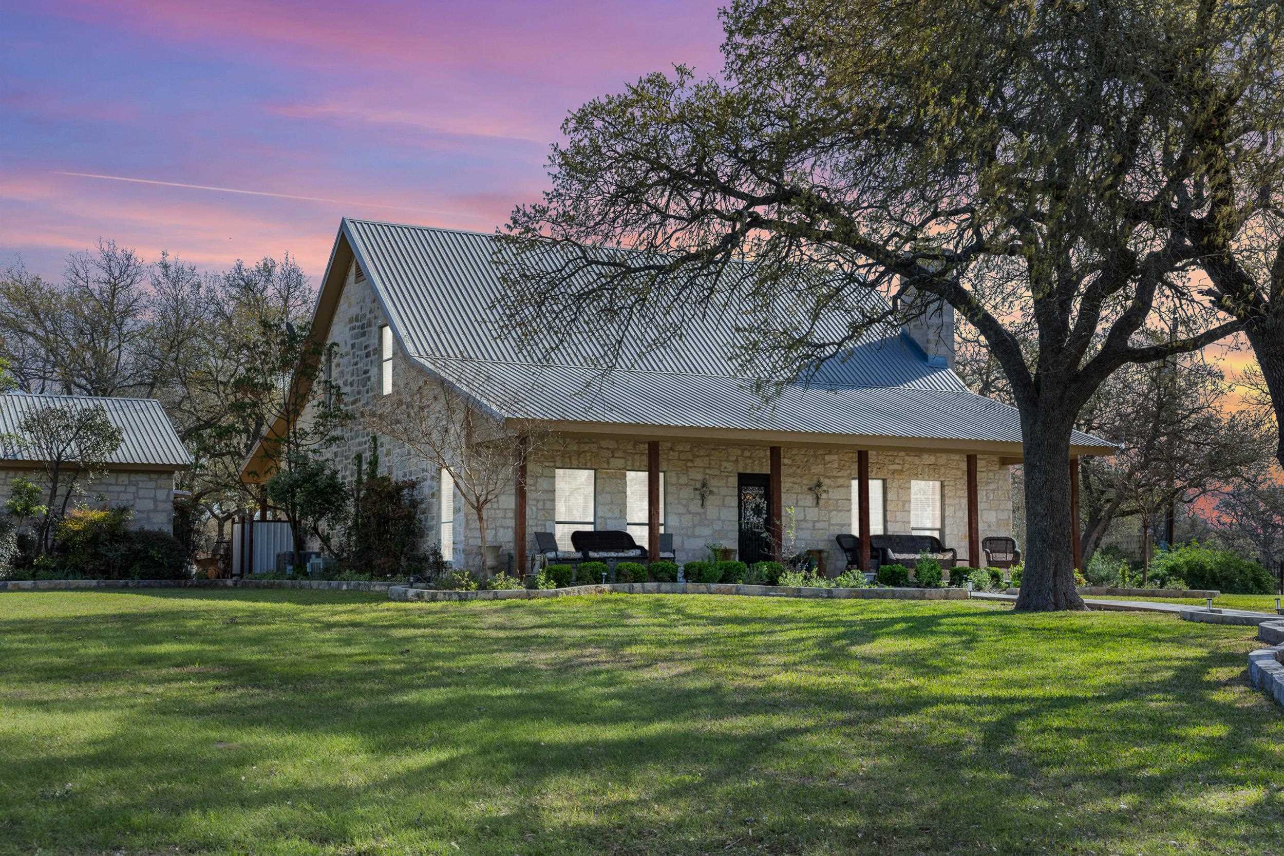 Stone house with a metal roof and covered porch beneath a large oak tree at dusk.