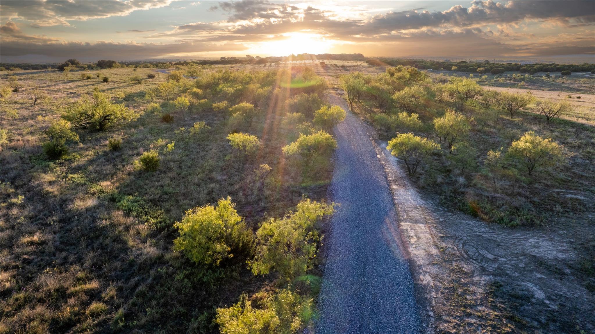 Aerial view of scrubland with a gravel road winding toward the golden sunset.