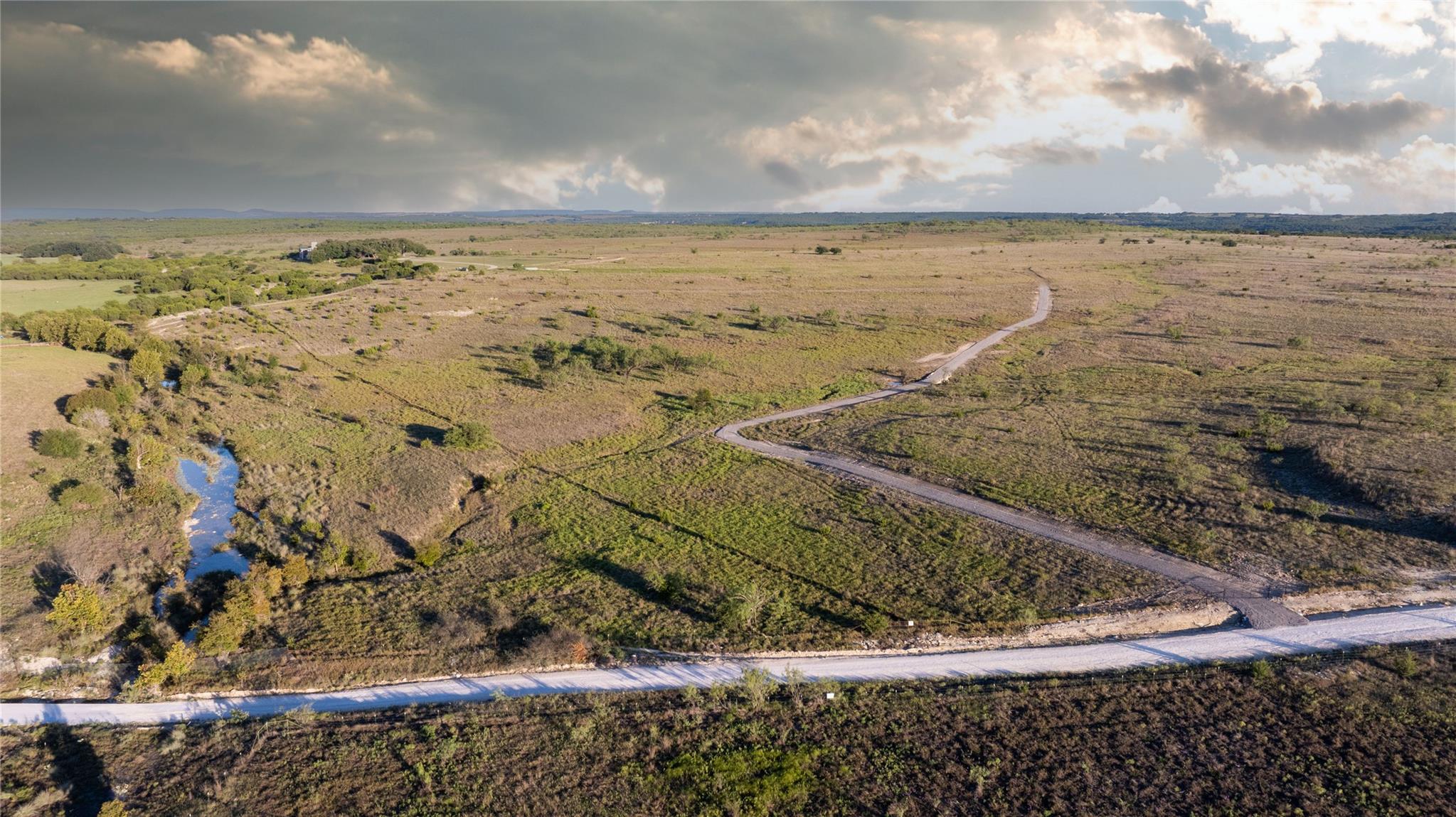 Aerial view of scrubland with a winding access road, paved road, and tree-lined creek.
