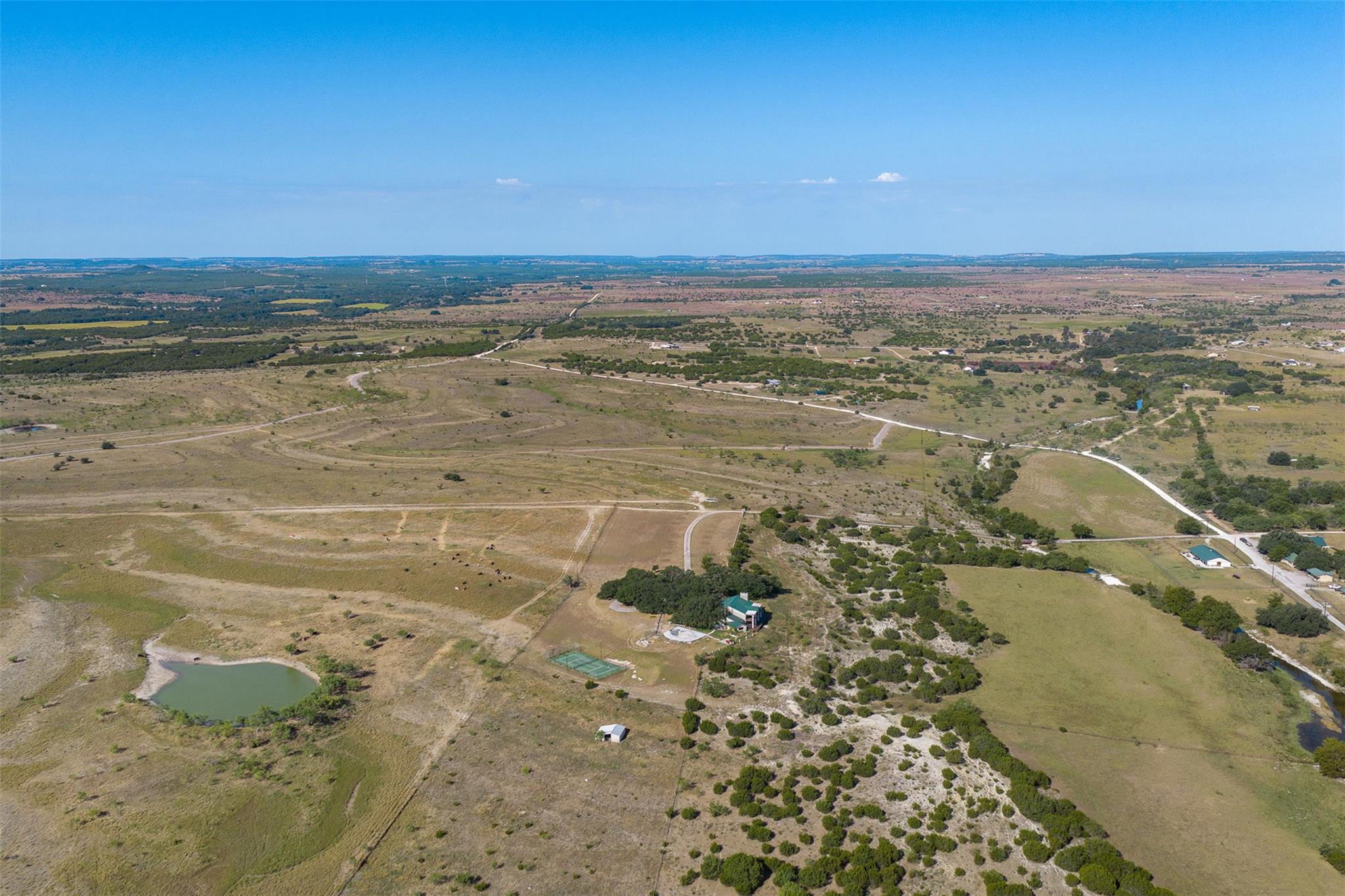 Aerial view of open rural land with a pond, scattered shrubs, dirt roads, and a few buildings.