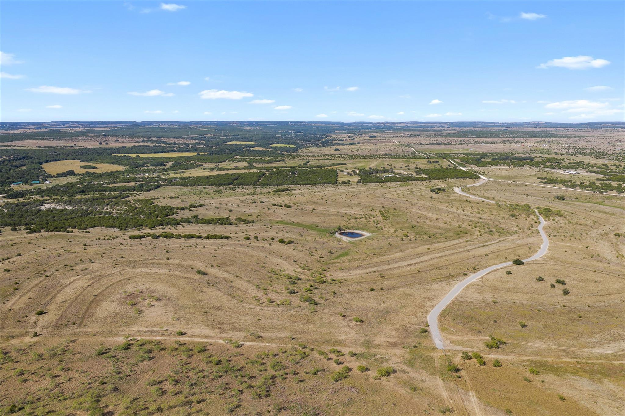 Aerial view of dry open land with scattered brush, a small pond, and a winding paved road.