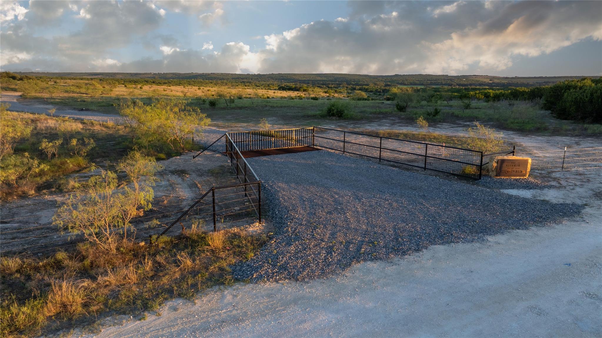 Rural lot with a gated gravel entrance, scrubland, and distant wooded hills in evening light.
