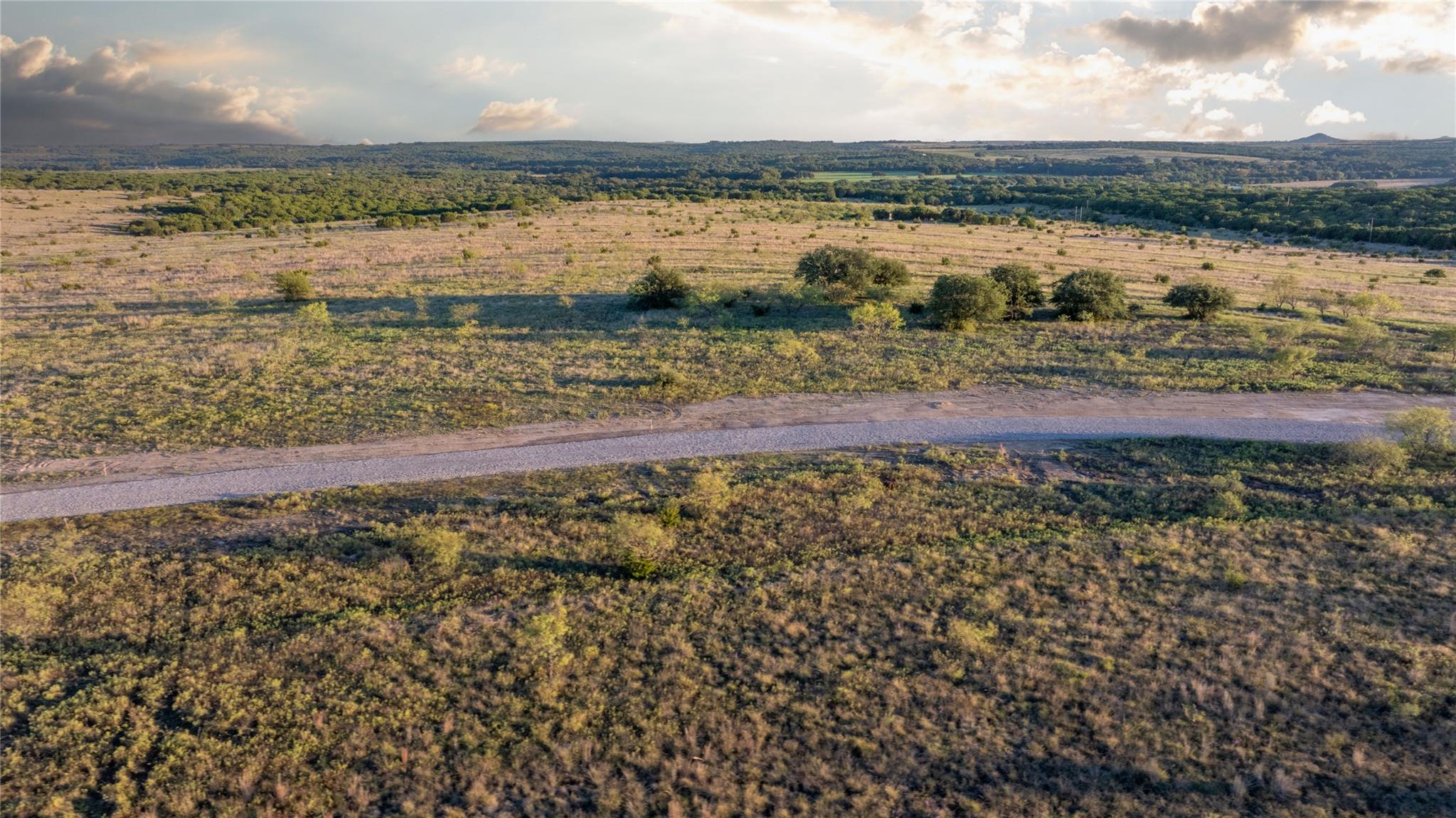 Aerial view of vacant scrubland crossed by a gravel road, with wooded hills in the distance.