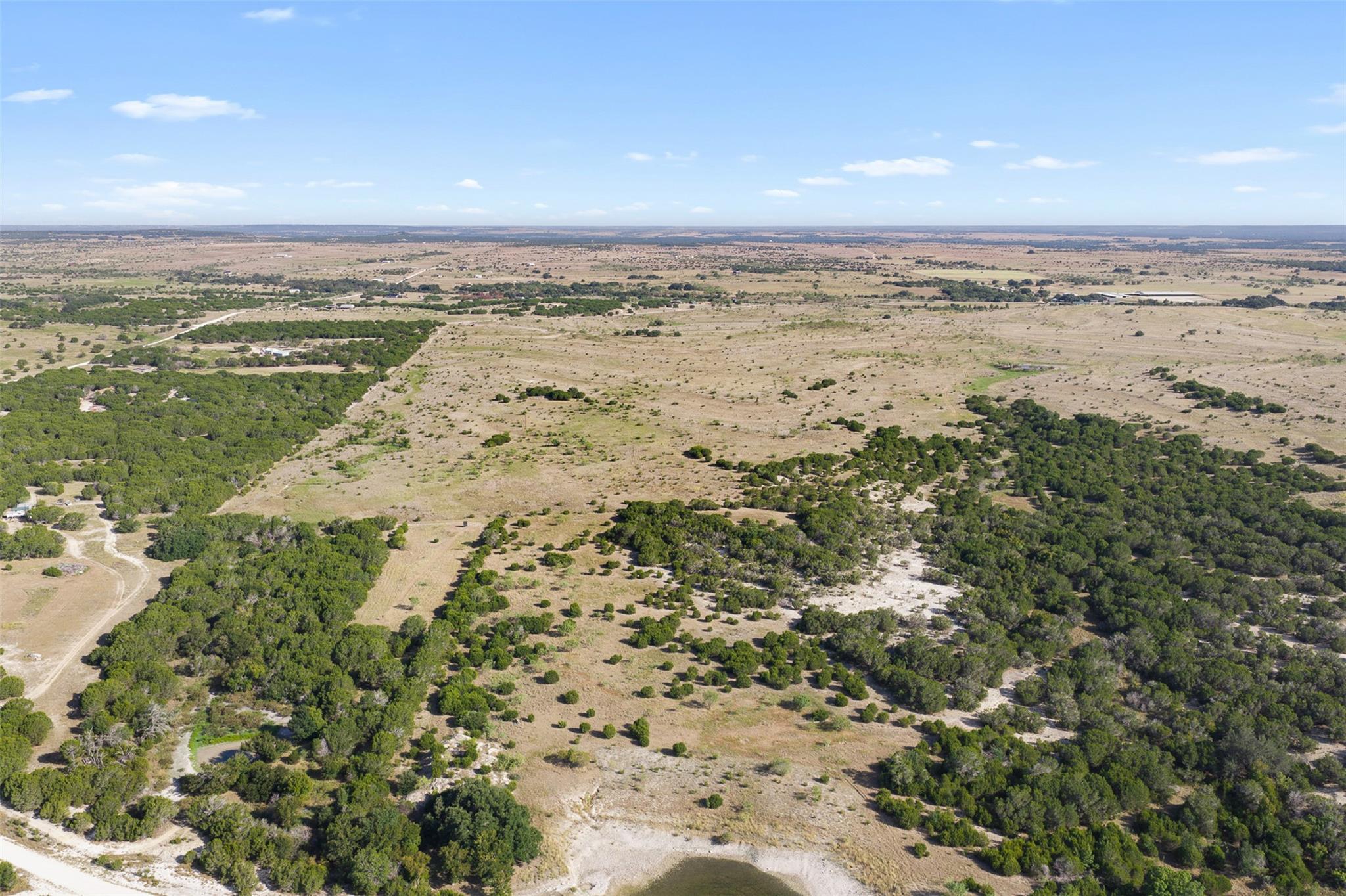 Aerial view of open rural land with scattered trees, dirt tracks, and a small pond.