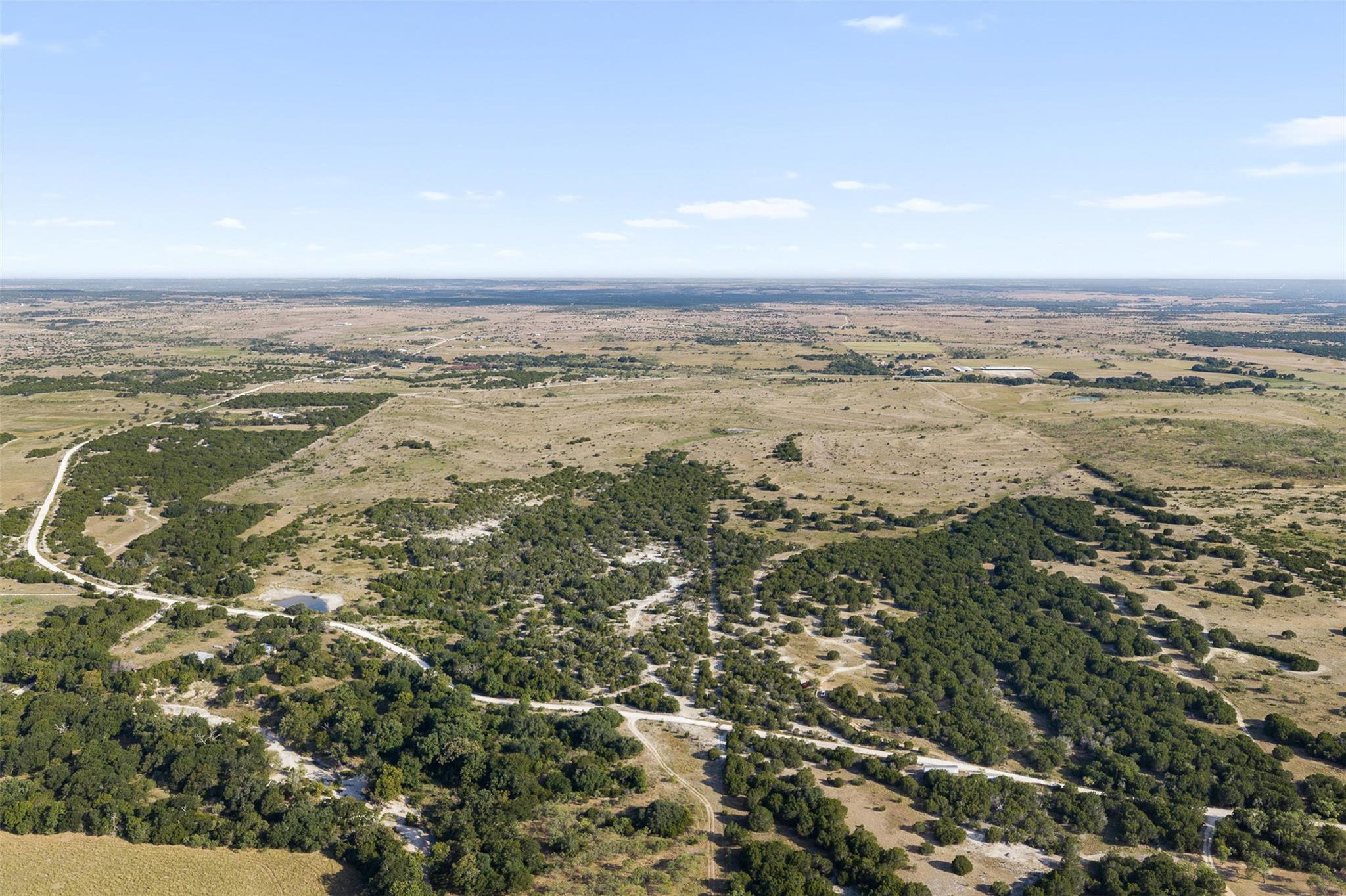 Aerial view of rural acreage with winding roads, scrubby trees, open grassland, and a small pond.