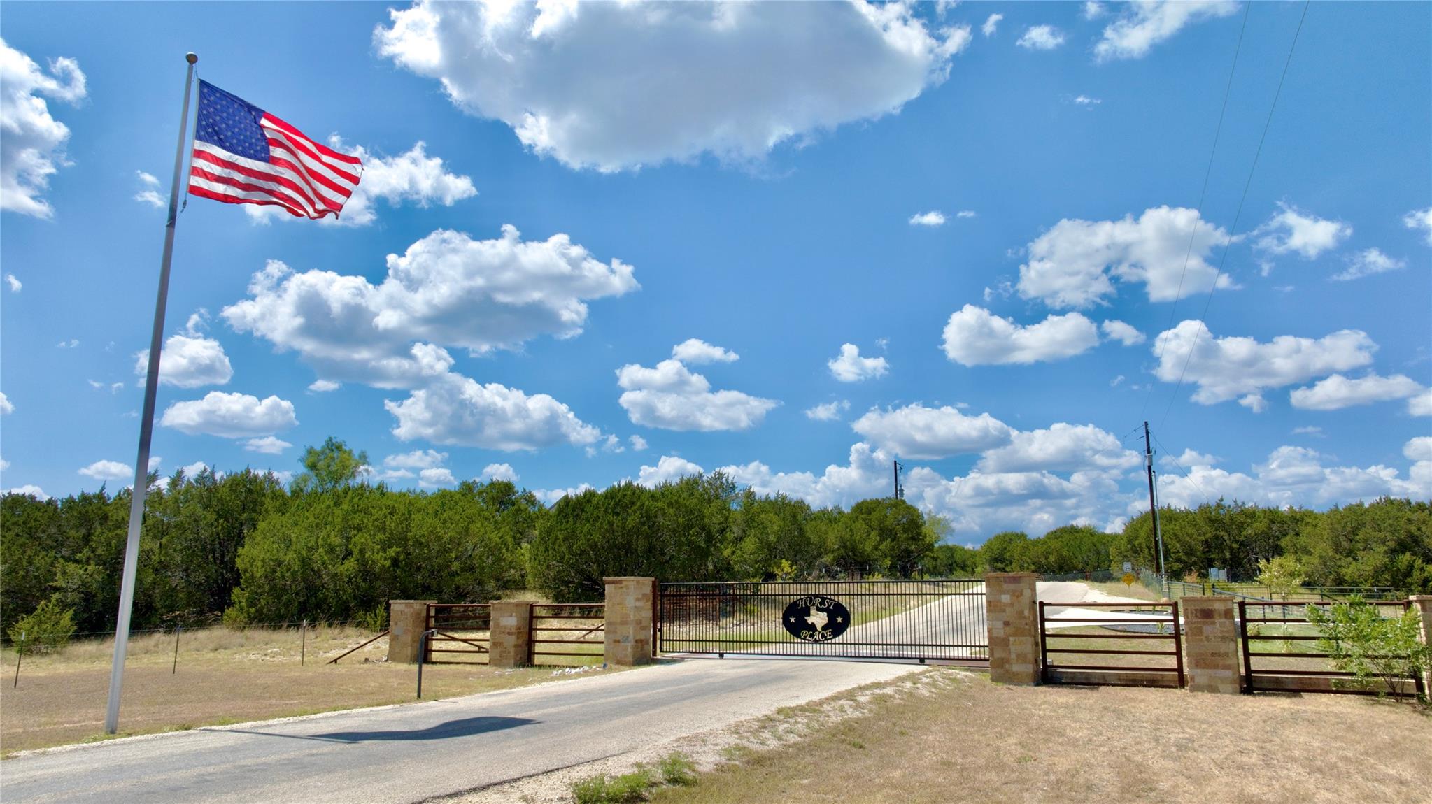 Gated property entrance with stone pillars, an American flag, and a wooded driveway.