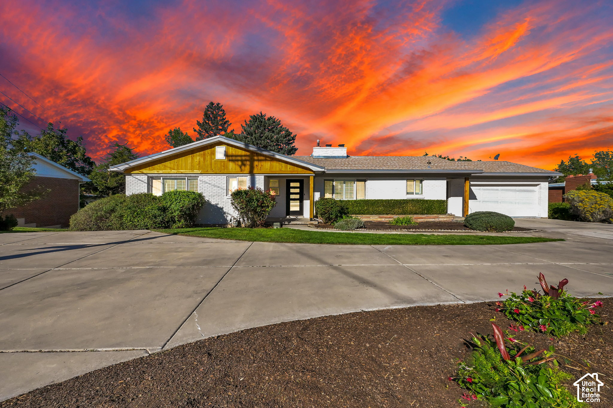 Single-story white house with wood siding, attached garage, and landscaped front yard beneath a vivid sunset.