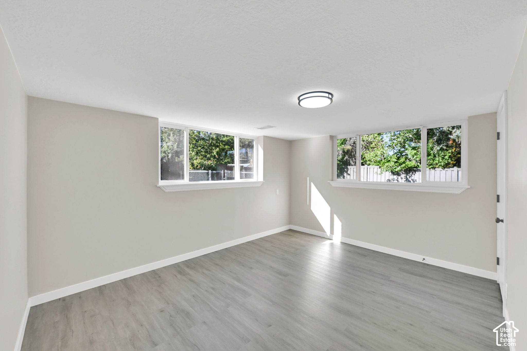 Vacant room with gray wood-look flooring and wide windows overlooking leafy trees.