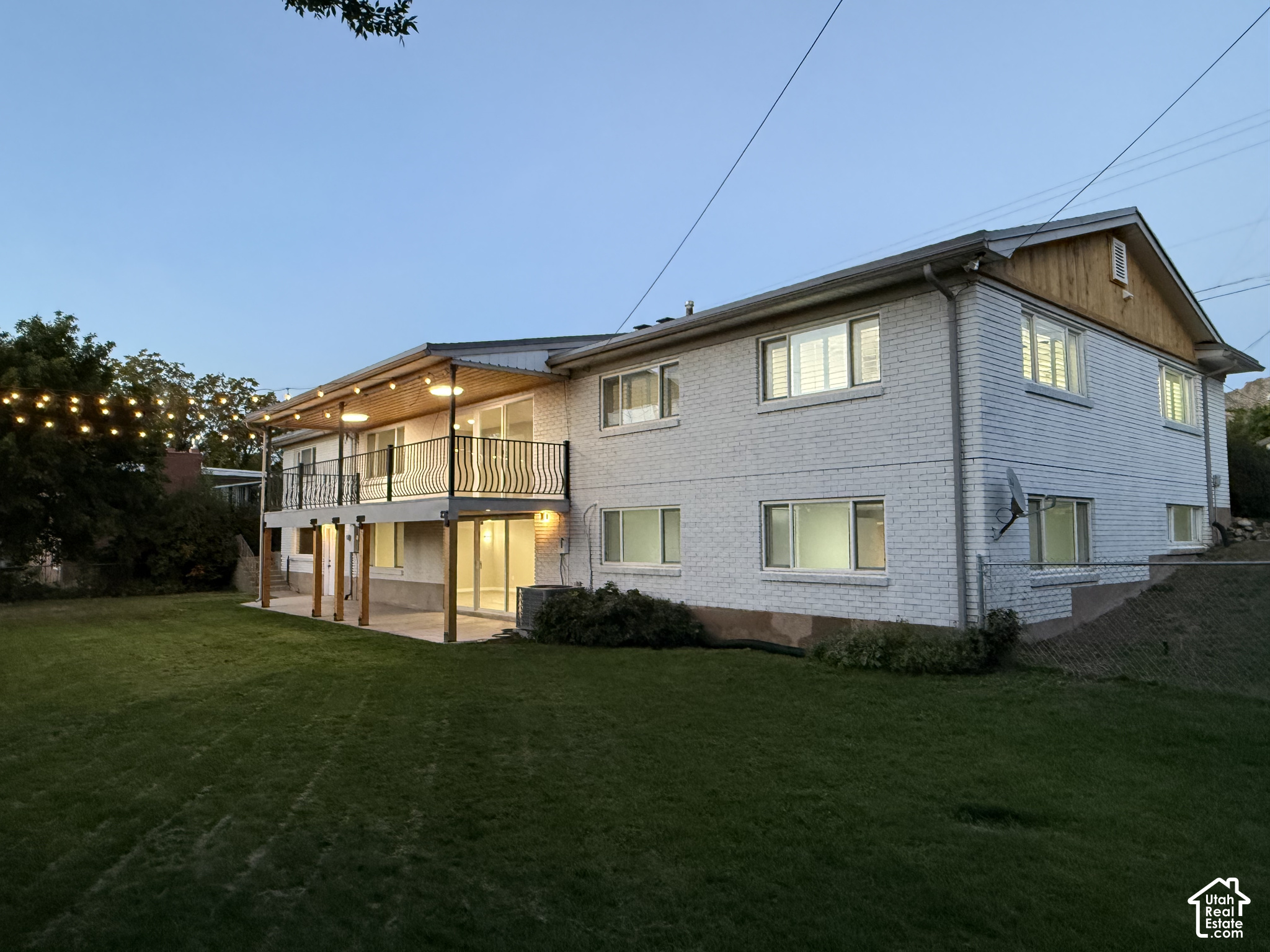 Two-story white brick home with a covered deck and string lights overlooking a backyard lawn at dusk.
