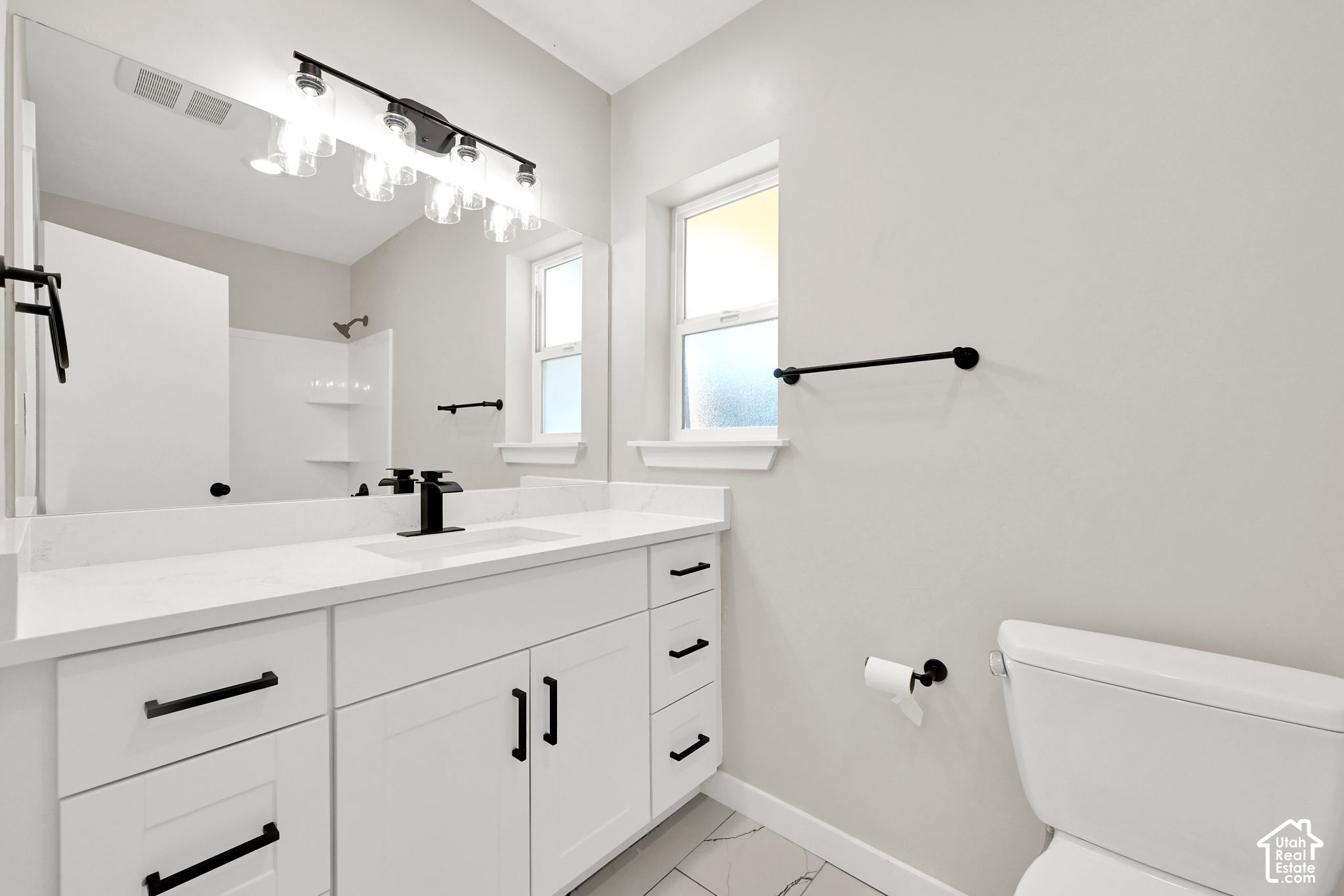 Modern bathroom with a white vanity, black fixtures, two windows, and gray veined tile flooring.