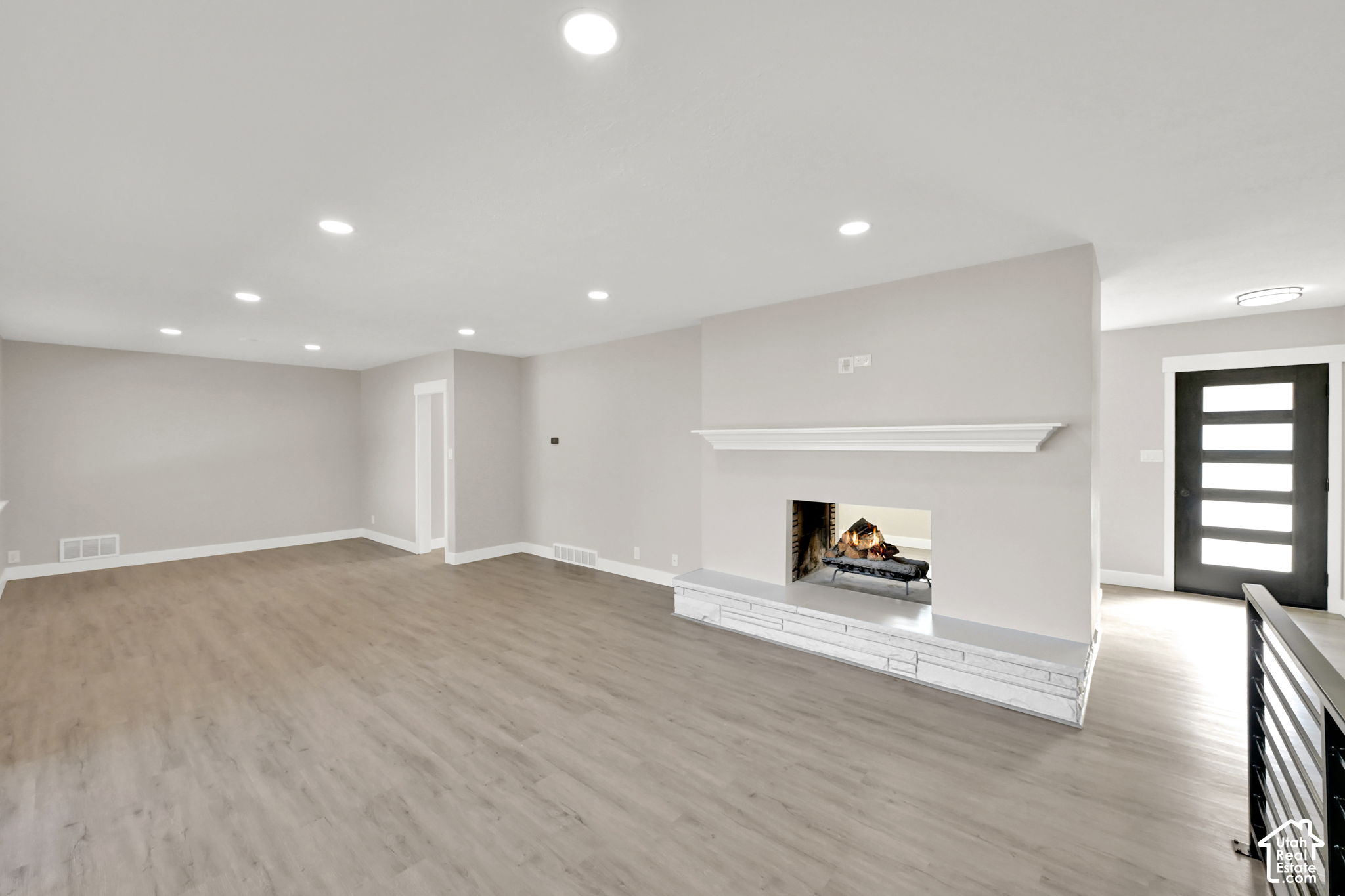 Empty living room with light wood floors, a white fireplace mantel, and a black glass-panel front door.