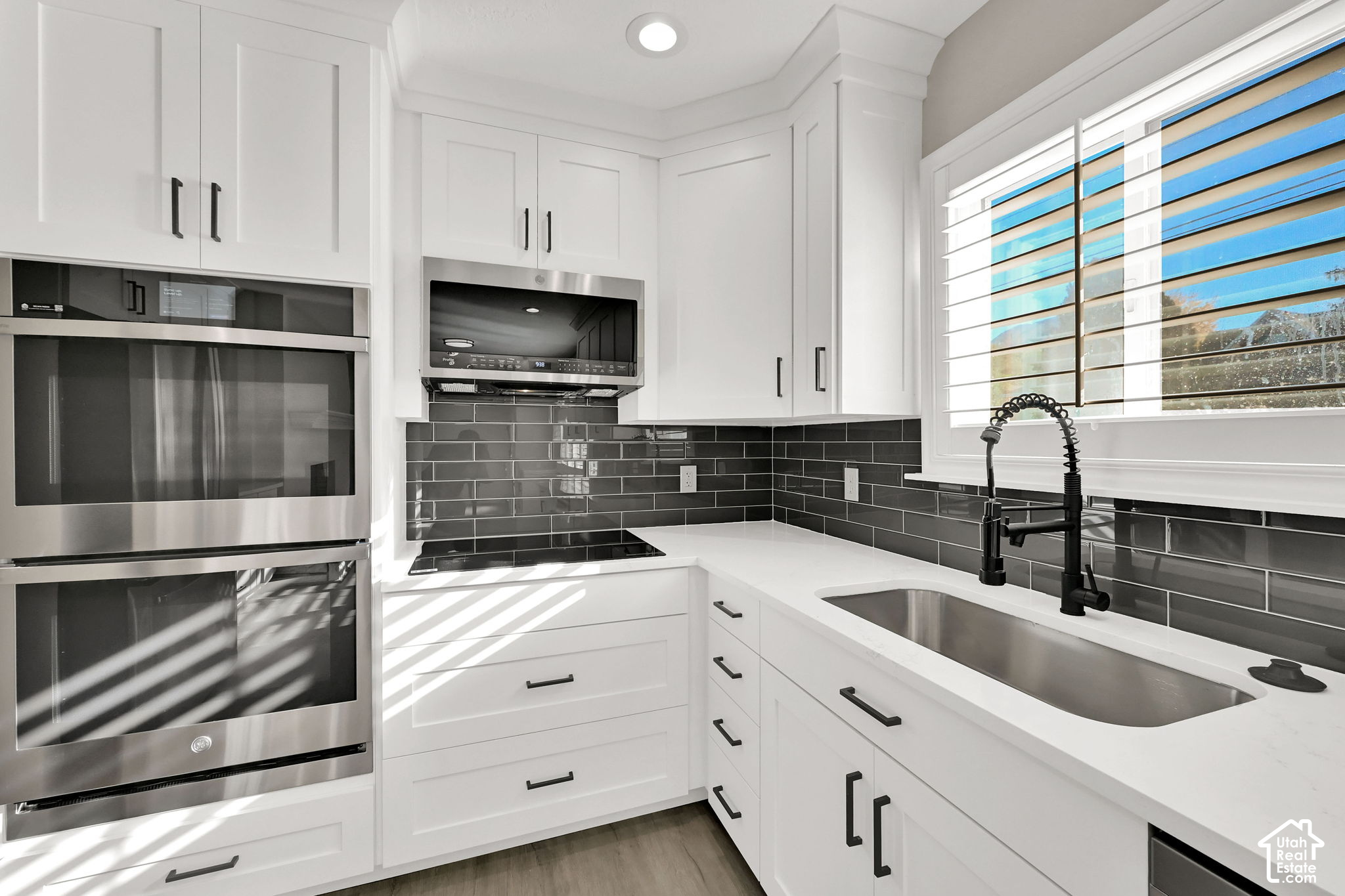 Modern white kitchen with double ovens, charcoal tile backsplash, and a large sink beneath a bright window.