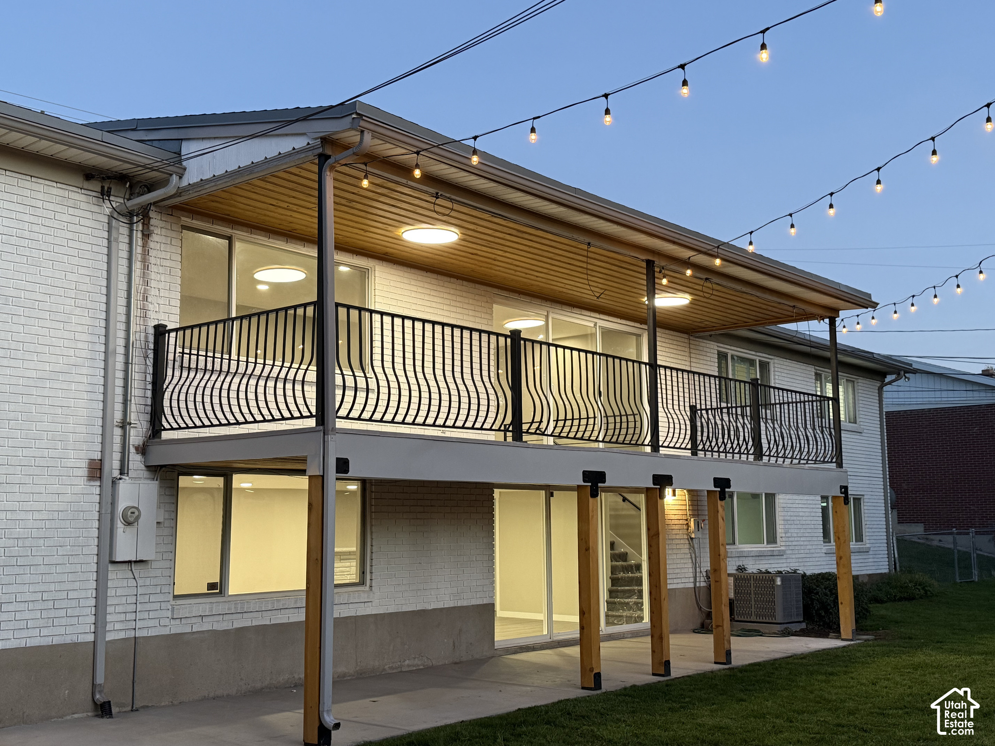 White brick home with an elevated deck, covered patio, and glowing string lights at dusk.