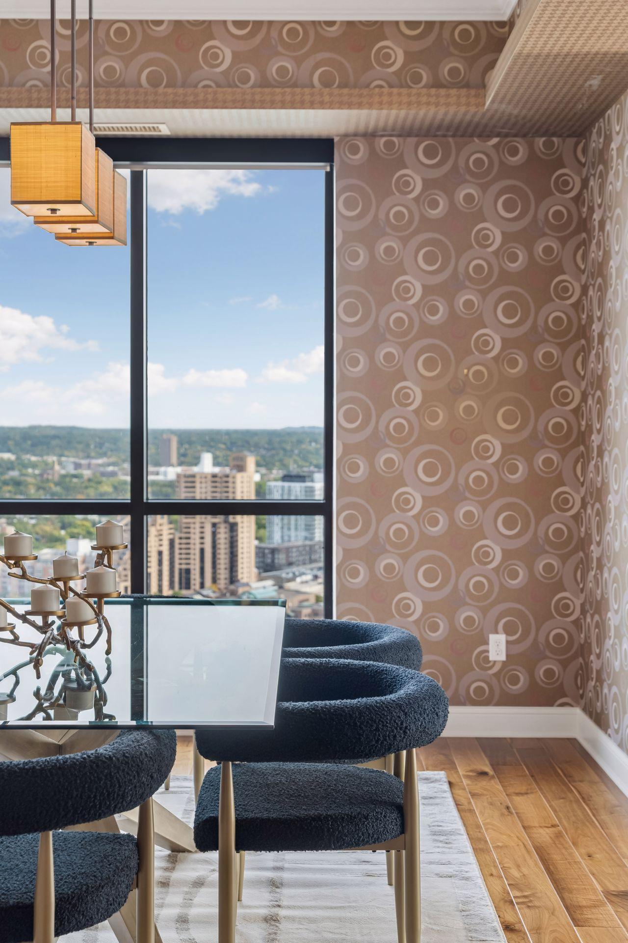 Dining area with dark upholstered chairs and patterned walls, overlooking the city through large windows.