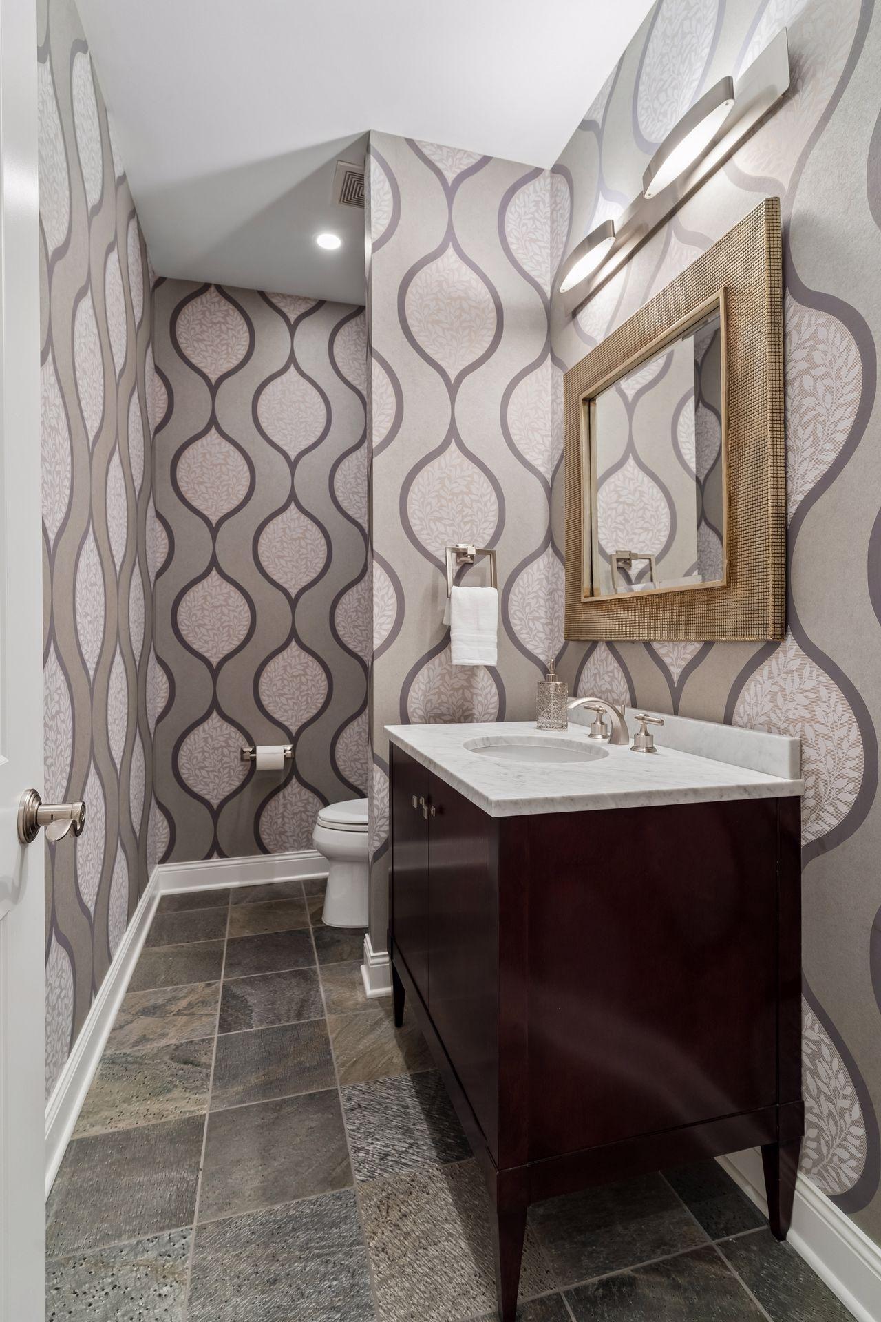 Powder room with patterned wallpaper, a dark wood vanity, marble countertop, and framed mirror.