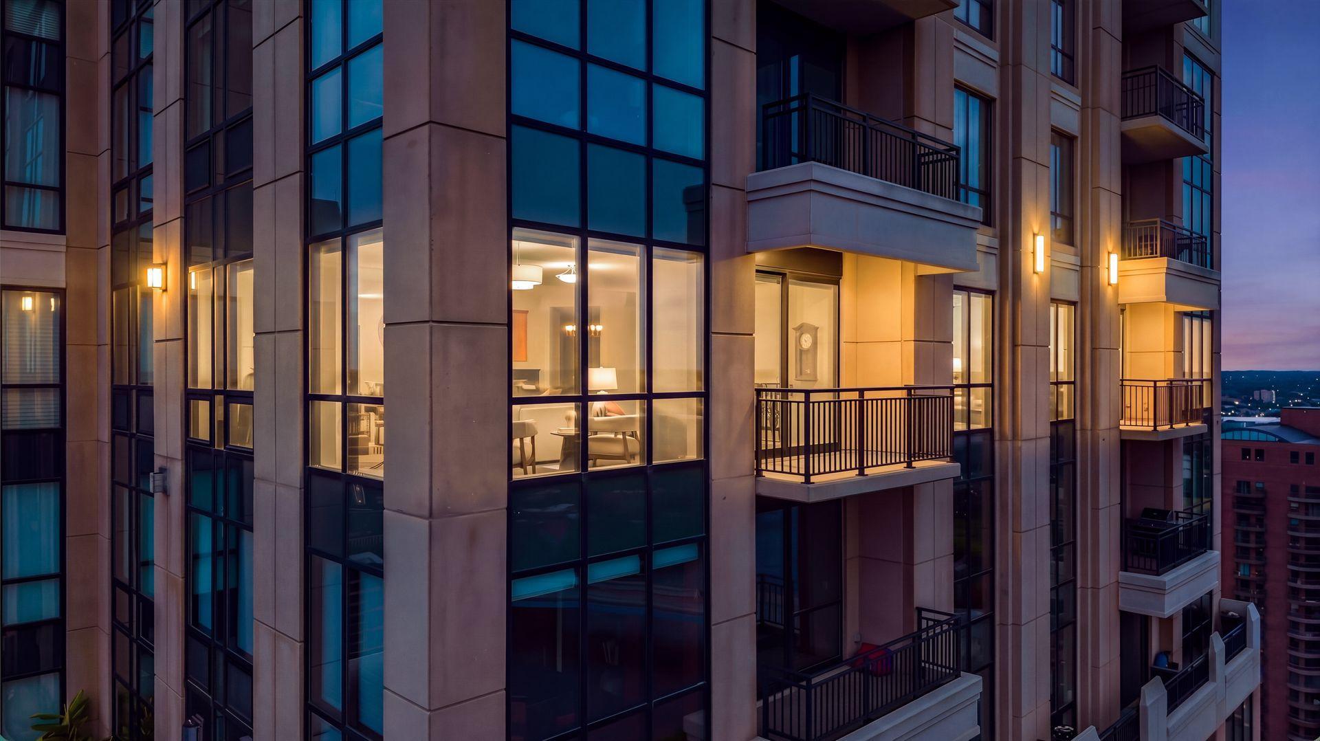 Modern condominium tower at dusk with glass windows, illuminated interiors, and balconies.