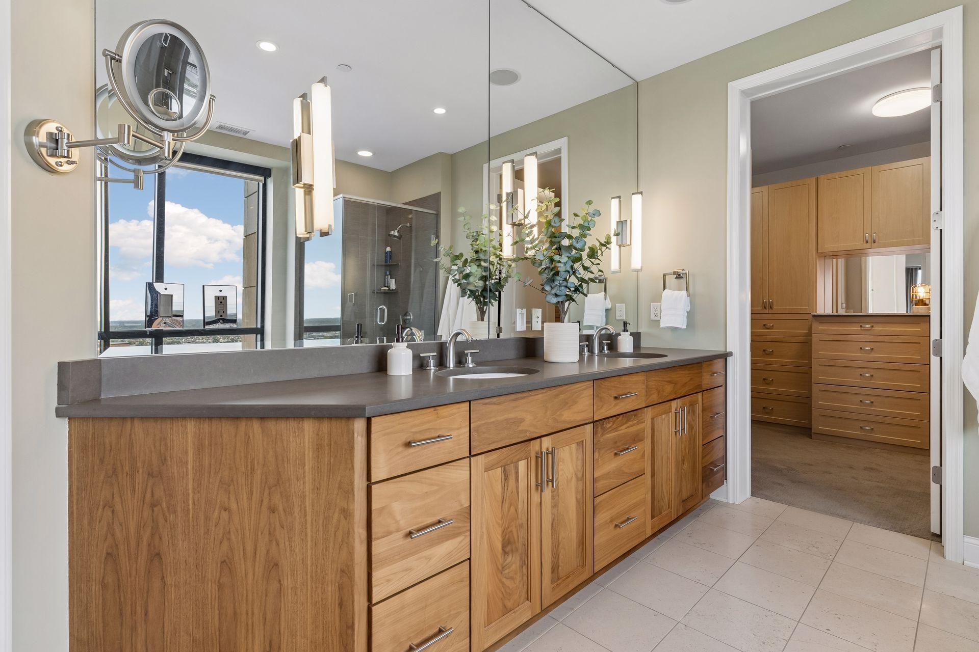 Modern bathroom with a long wood double vanity, mirrored wall, glass shower, and doorway to a walk-in closet.