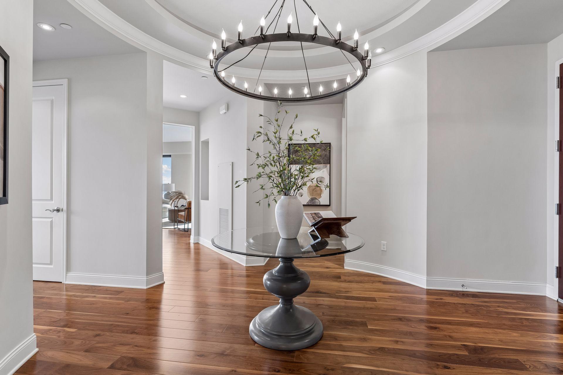 Dining area with a glass-top table, leafy centerpiece, circular chandelier, and hardwood floors.