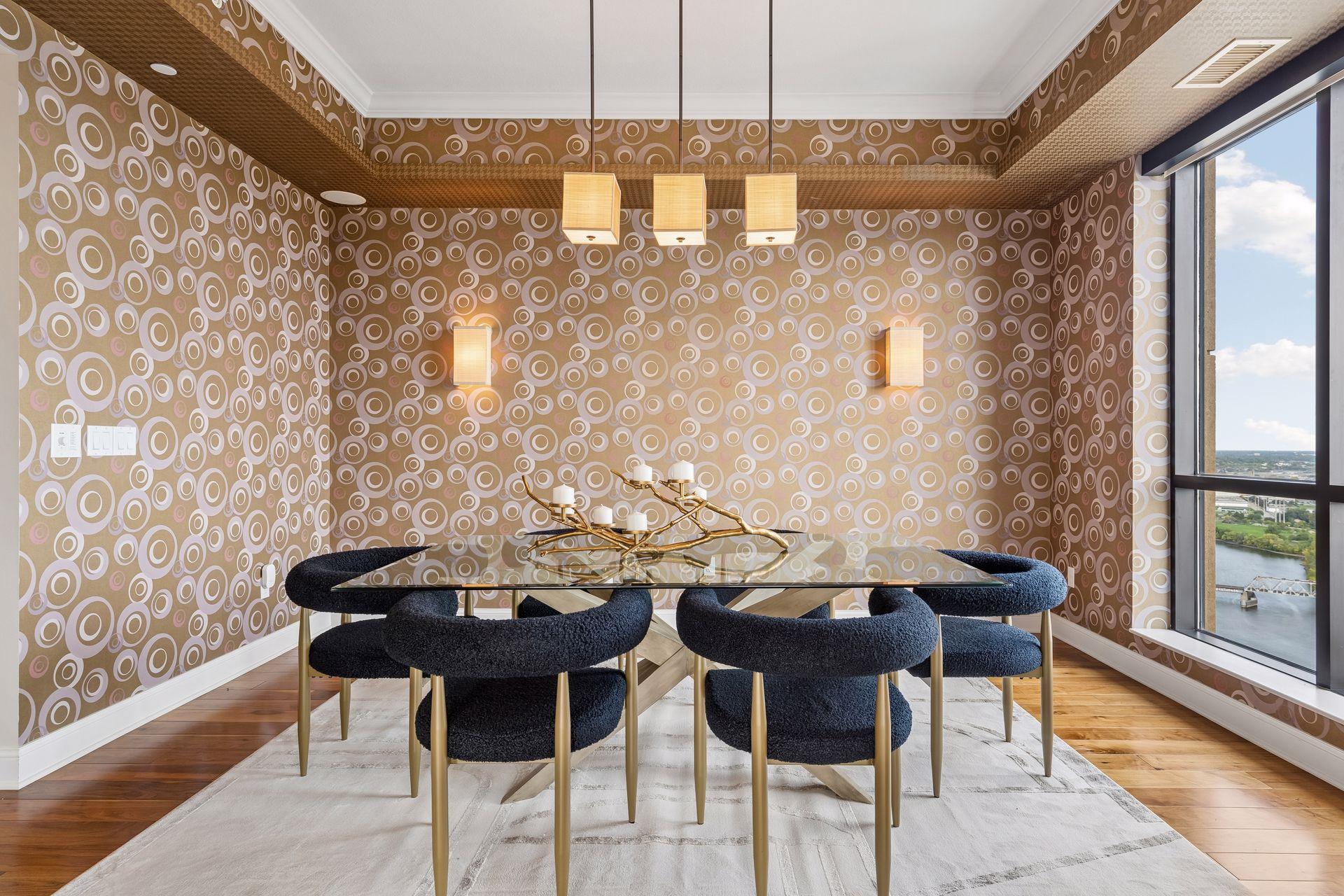 Dining room with circular-patterned wallpaper, a glass table, dark chairs, and river views through tall windows.