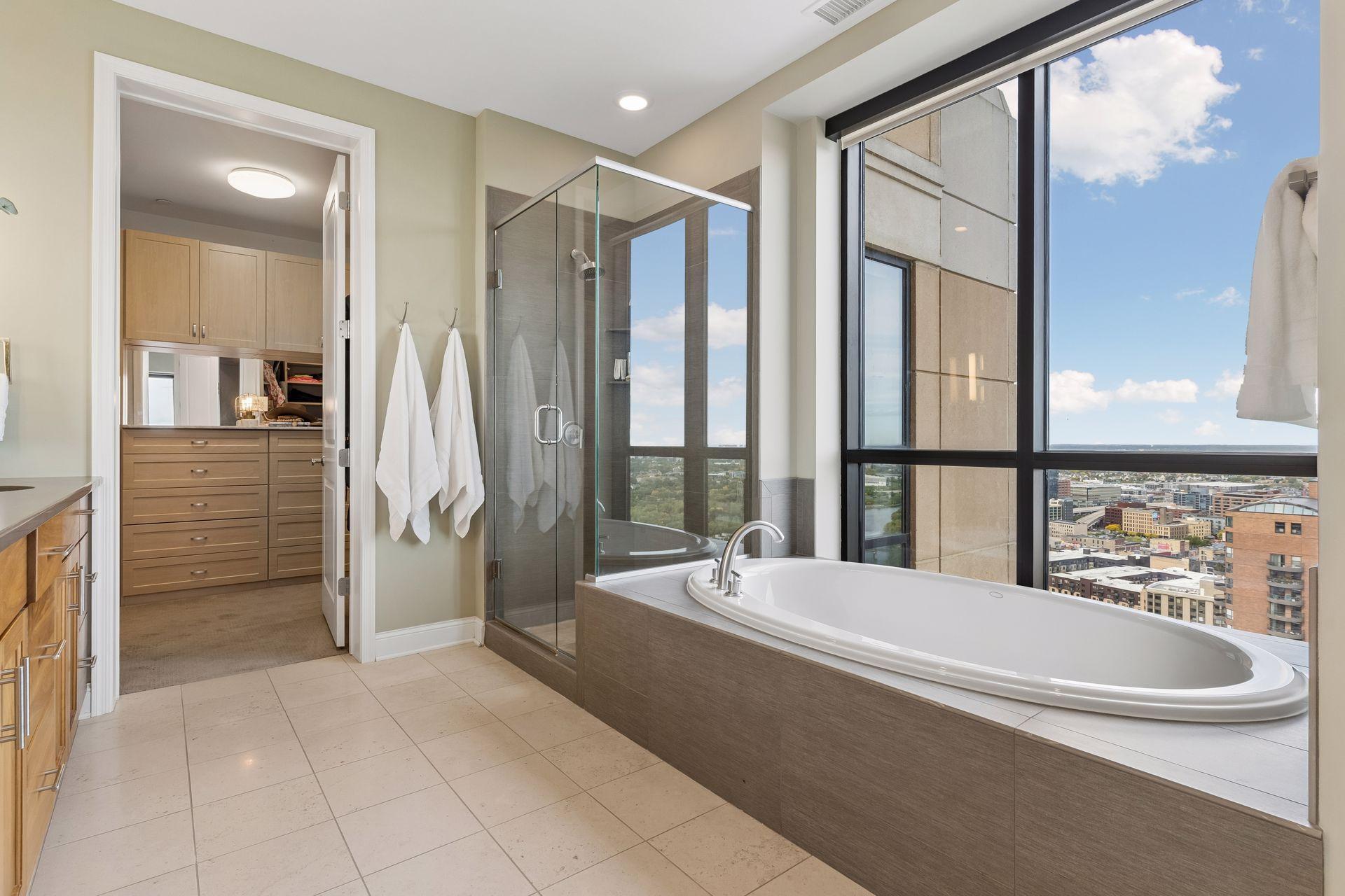 Condo bathroom with a glass shower, large soaking tub, and city views through floor-to-ceiling windows.