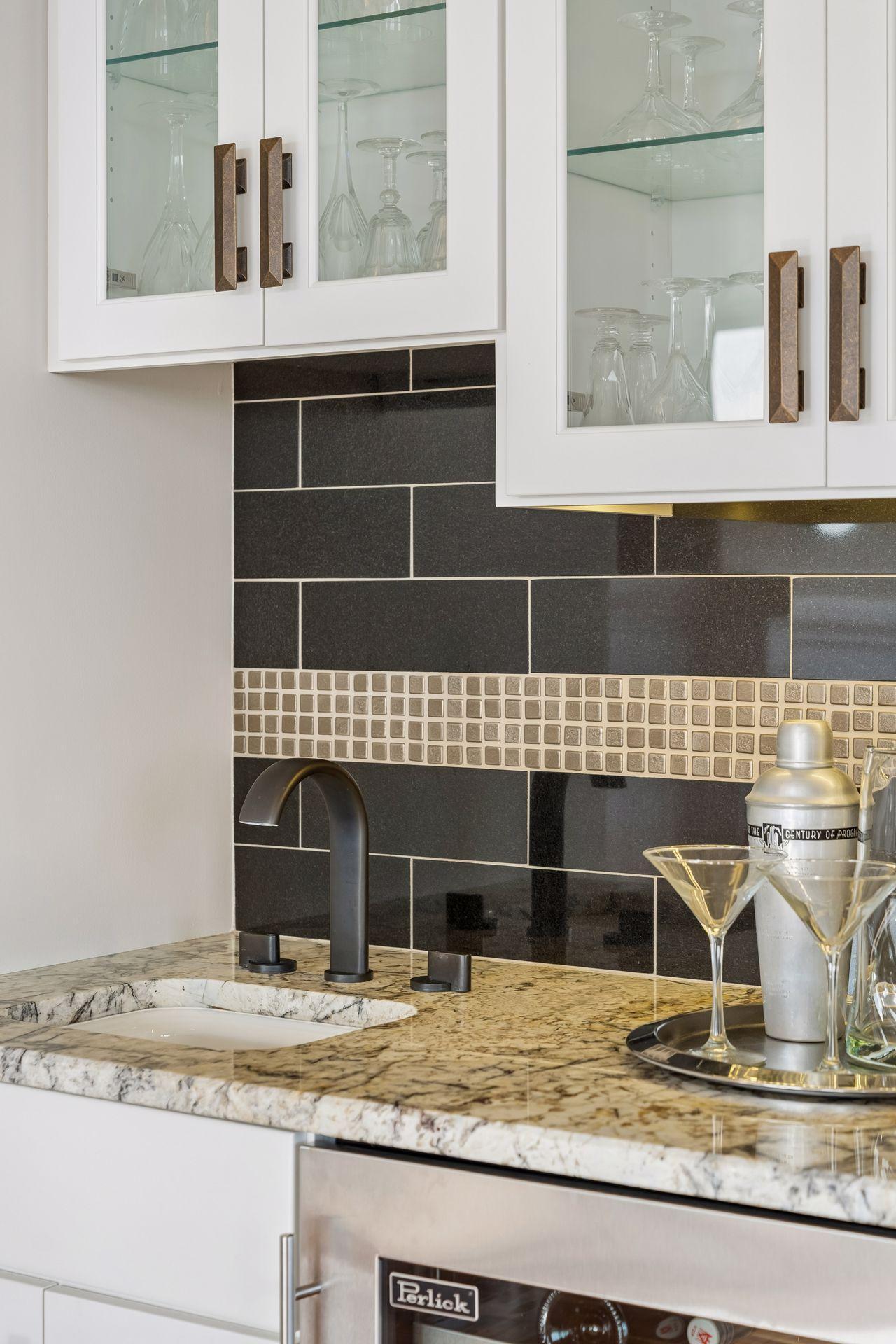 Kitchen wet bar with glass-front cabinets, dark tile backsplash, veined stone counter, and matte black faucet.