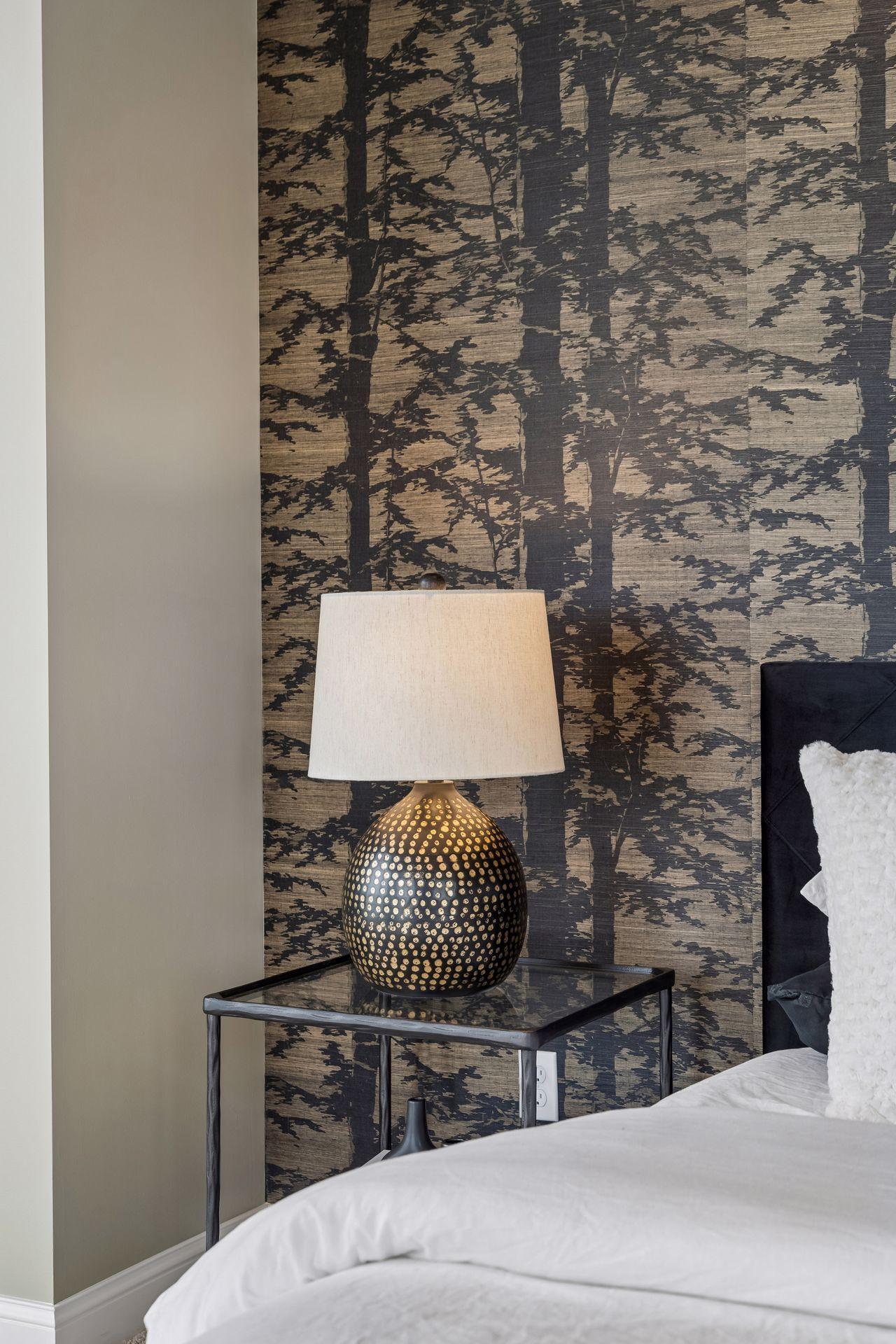 Bedroom corner with patterned accent wall, decorative lamp, glass bedside table, and white bed.